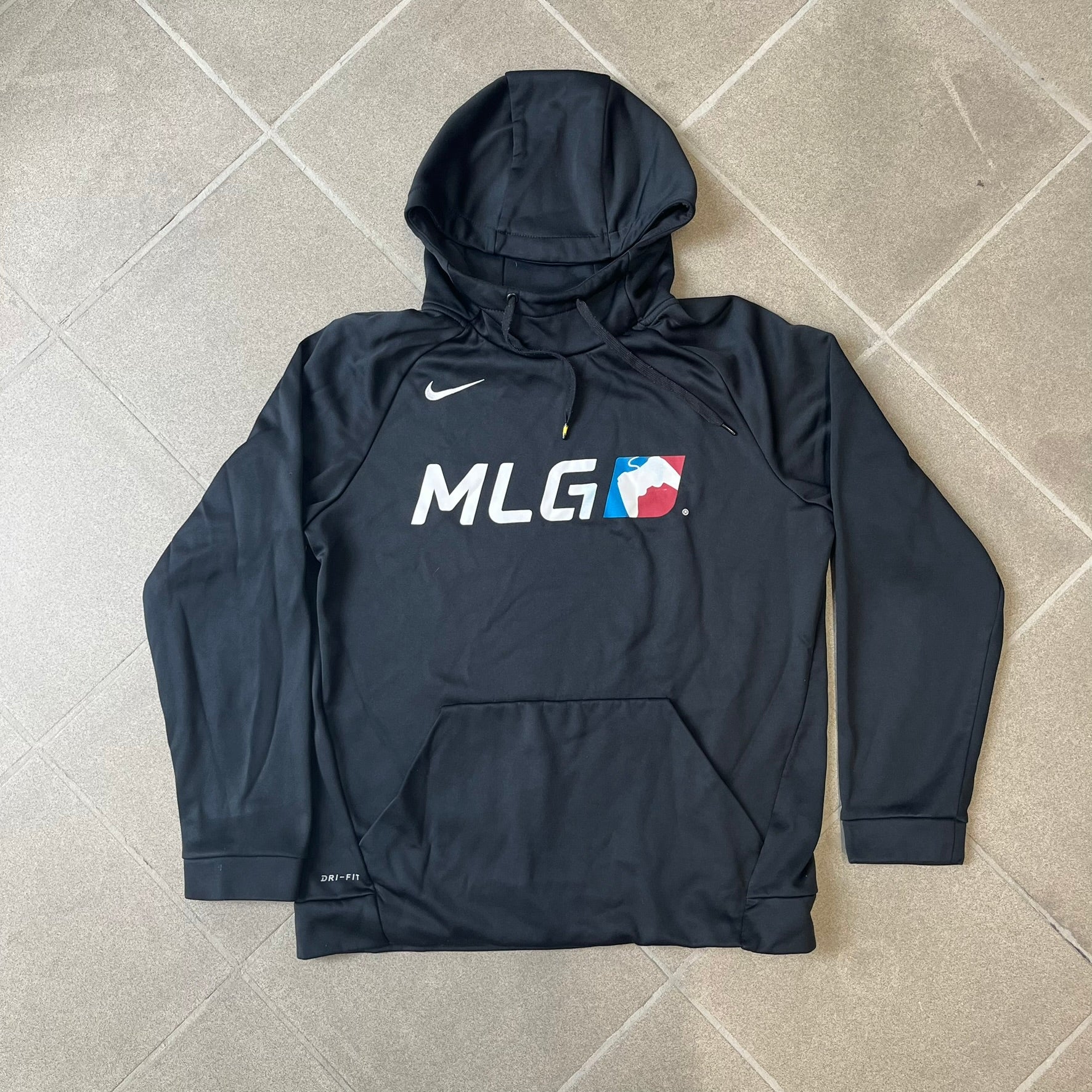 Nike MLG (Major League Gaming) Black Hoodie is a performance hoodie made from 100% polyester with Nike Dri-FIT technology. It features a kangaroo pocket, a Nike Swoosh on the upper right chest, and a large Major League Gaming (MLG) logo across the center with the iconic red and blue gamepad graphic. The hood has a raised front design that extends to cover the mouth.