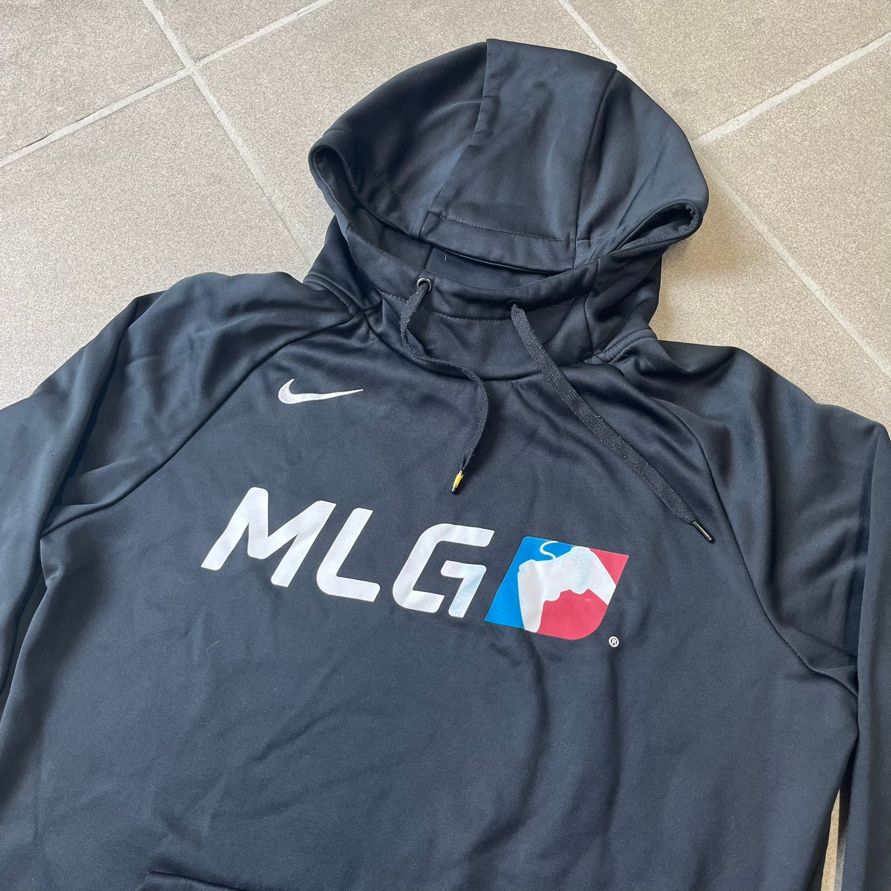 Nike MLG (Major League Gaming) Black Hoodie is a performance hoodie made from 100% polyester with Nike Dri-FIT technology. It features a kangaroo pocket, a Nike Swoosh on the upper right chest, and a large Major League Gaming (MLG) logo across the center with the iconic red and blue gamepad graphic. The hood has a raised front design that extends to cover the mouth.