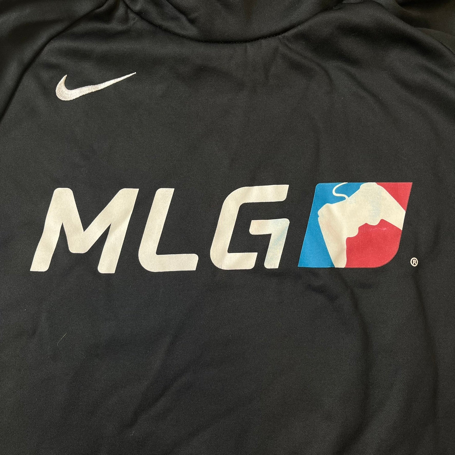 Nike MLG (Major League Gaming) Black Hoodie is a performance hoodie made from 100% polyester with Nike Dri-FIT technology. It features a kangaroo pocket, a Nike Swoosh on the upper right chest, and a large Major League Gaming (MLG) logo across the center with the iconic red and blue gamepad graphic. The hood has a raised front design that extends to cover the mouth.