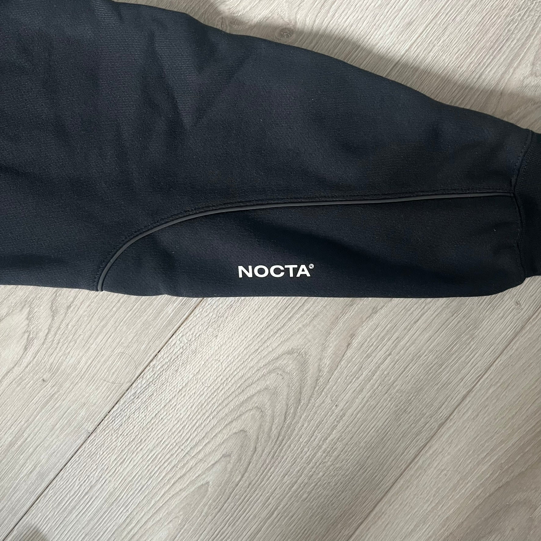 Nike x Nocta Fleece CS Hoodie in Black combines Drake’s refined streetwear vision with Nike’s premium craftsmanship. Made from an ultra-soft fleece blend, it features reflective piping, a silicone Swoosh logo and discreet NOCTA branding. With an adjustable hood, kangaroo pocket, and hidden zip pocket, this piece balances luxury comfort and technical precision.
