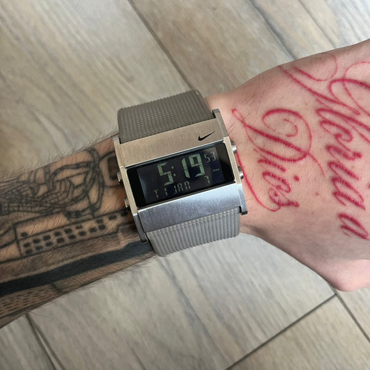 Nike Oregon Series Square Black Face Digital Watch WA0036-201 is a digital wristwatch featuring a rectangular display with a stainless steel frame and a light grey rubber strap with stainless steel clasp. The watch includes four side buttons, backlight function, and displays digital numerals on a black screen, with a small Nike logo on the metal bezel. It is water resistant up to 100 meters, powered by fresh batteries, and comes with original tag and box