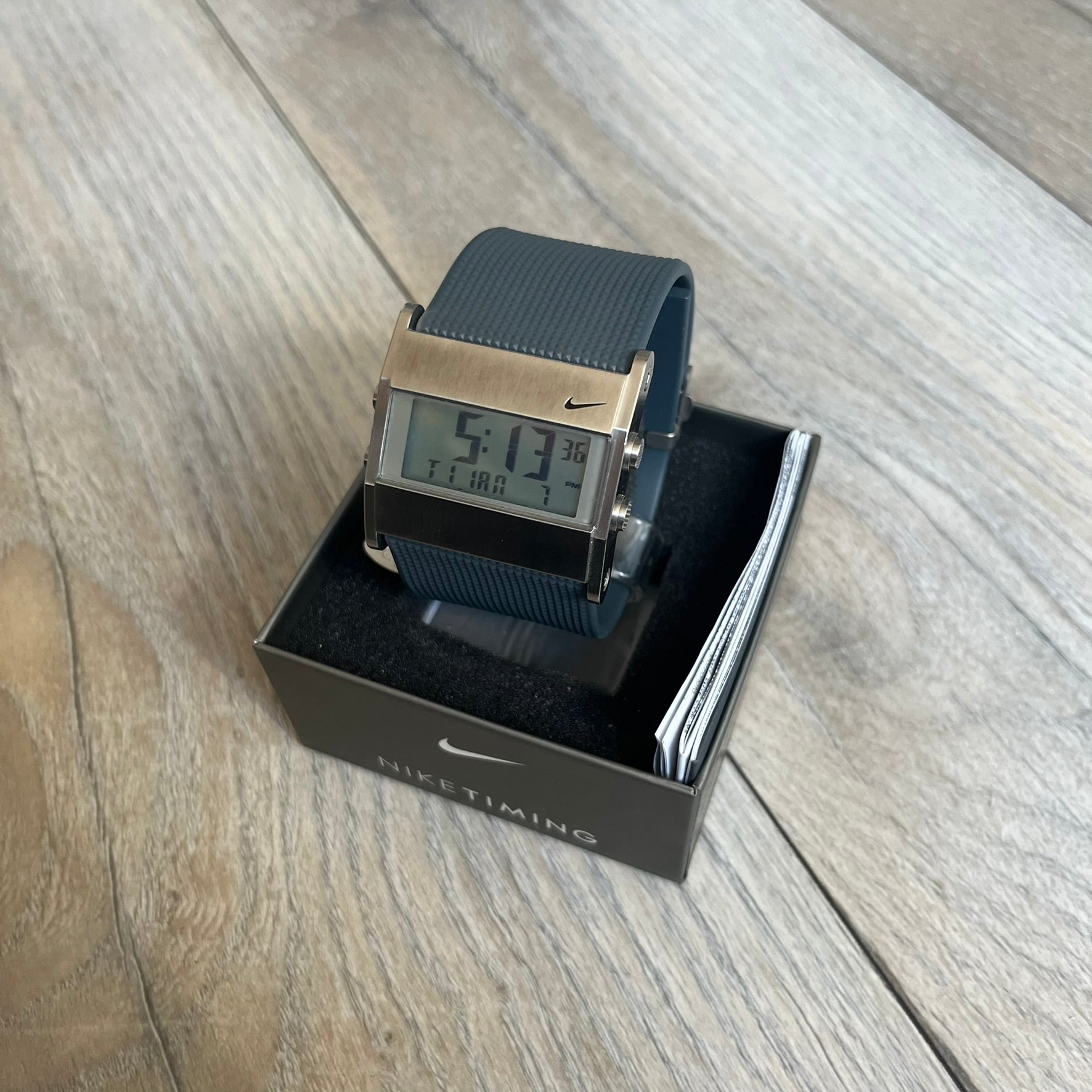 Nike Oregon Series Gray Digital Watch is a digital wristwatch featuring a rectangular display with a stainless steel frame and a dark grey rubber strap with stainless steel clasp. The watch includes four side buttons, backlight function, and displays black digital numerals, with a small Nike logo on the metal bezel. It is water resistant up to 100 meters, powered by fresh batteries, and comes with original tag and box.