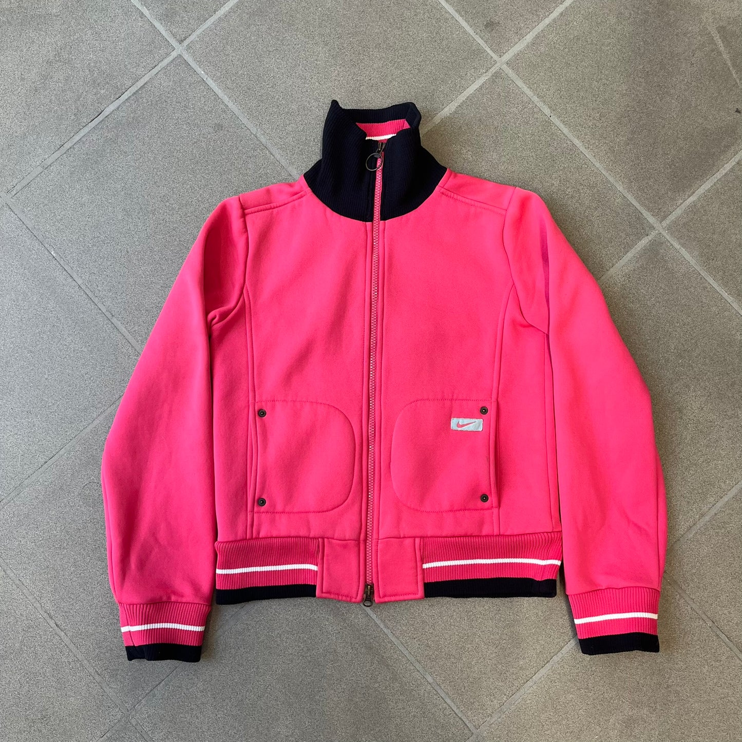 Nike Pink Women's Bomber-style Zip-up features a pink, heavyweight cotton construction with a tall black collar. It has a full front zipper with a circular zipper pull and two front Velcro pockets, one with a small Nike tag. The garment has a jacket-style construction.
