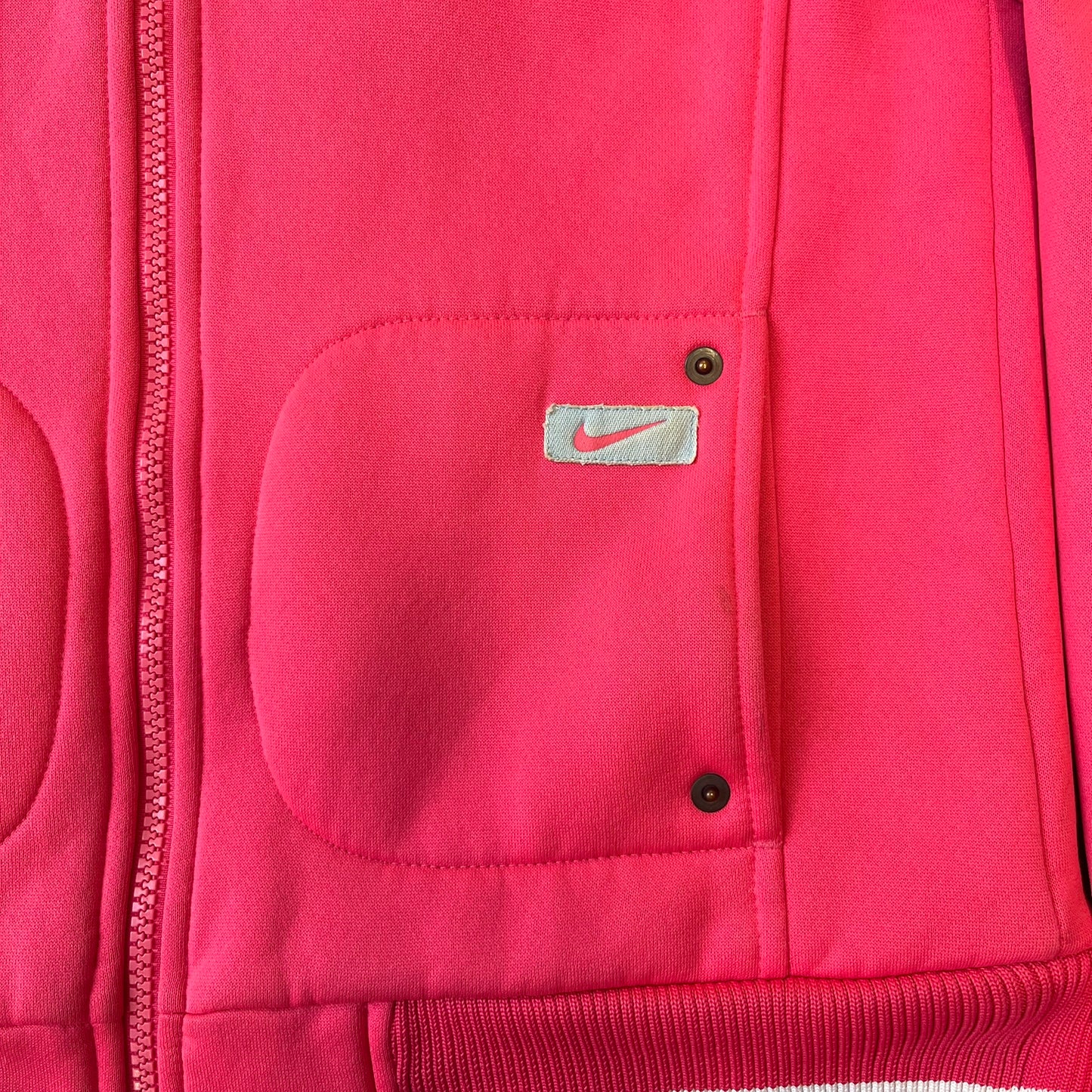 Nike Pink Women's Bomber-style Zip-up features a pink, heavyweight cotton construction with a tall black collar. It has a full front zipper with a circular zipper pull and two front Velcro pockets, one with a small Nike tag. The garment has a jacket-style construction.
