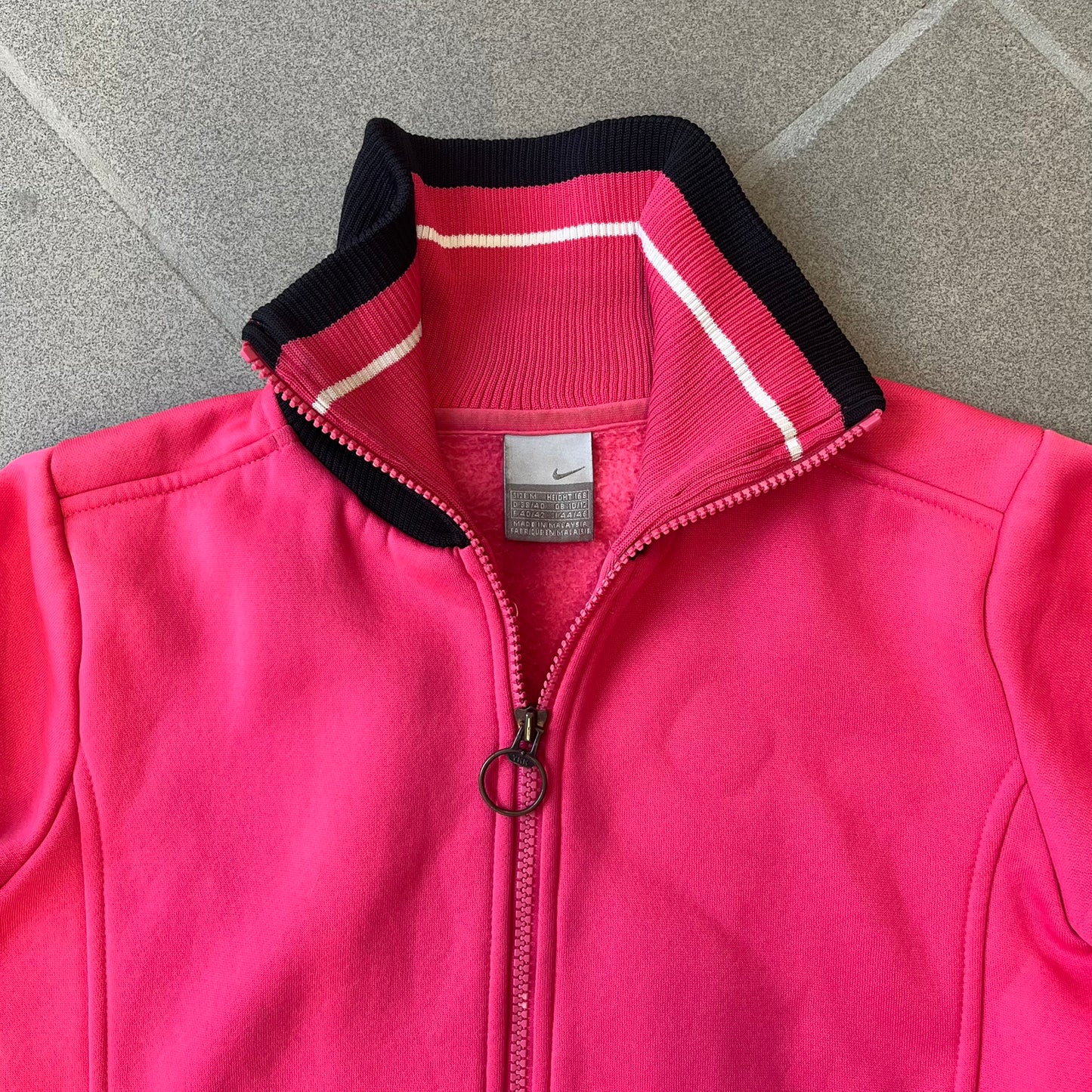 Nike Pink Women's Bomber-style Zip-up features a pink, heavyweight cotton construction with a tall black collar. It has a full front zipper with a circular zipper pull and two front Velcro pockets, one with a small Nike tag. The garment has a jacket-style construction.
