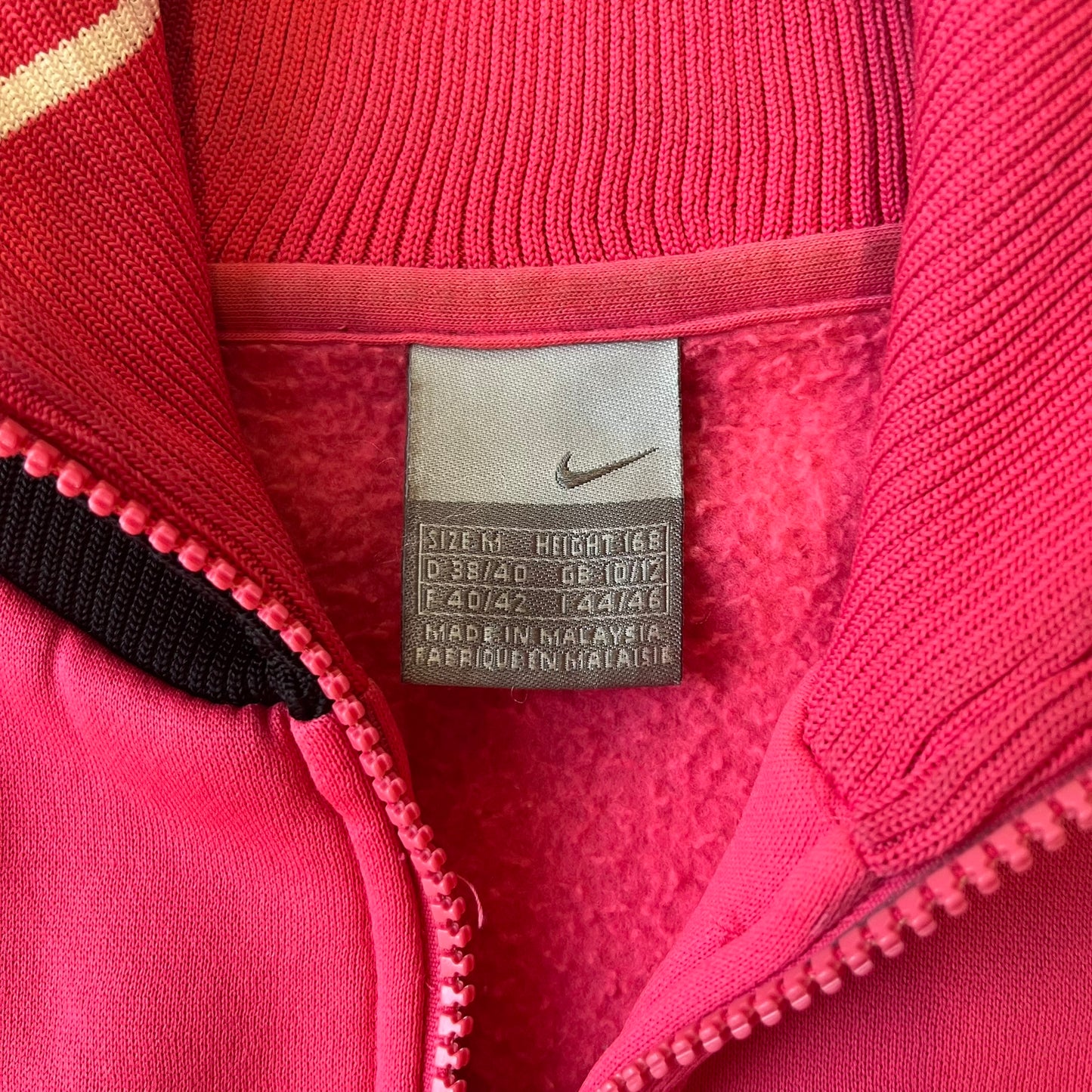 Nike Pink Women's Bomber-style Zip-up features a pink, heavyweight cotton construction with a tall black collar. It has a full front zipper with a circular zipper pull and two front Velcro pockets, one with a small Nike tag. The garment has a jacket-style construction.
