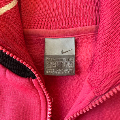 Nike Pink Women's Bomber-style Zip-up features a pink, heavyweight cotton construction with a tall black collar. It has a full front zipper with a circular zipper pull and two front Velcro pockets, one with a small Nike tag. The garment has a jacket-style construction.
