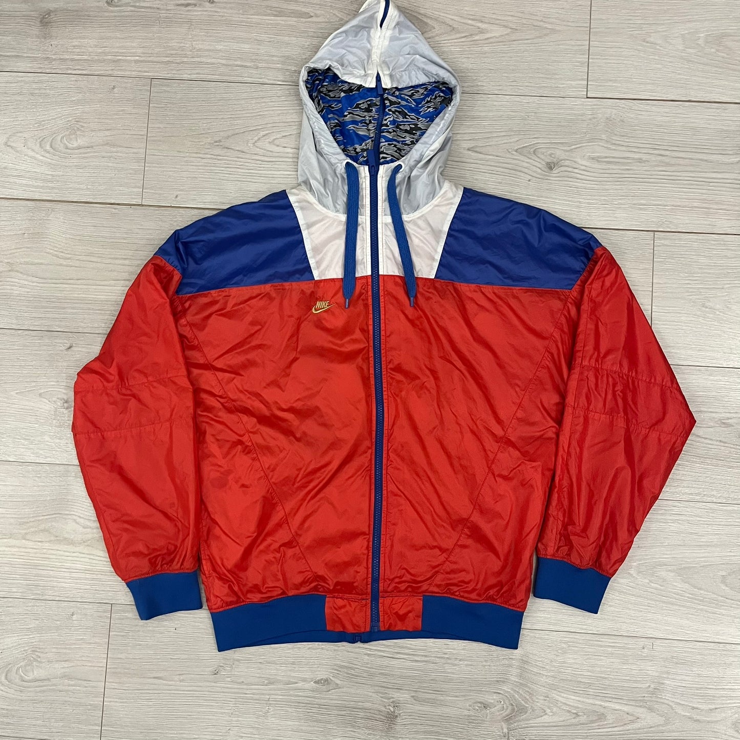 Nike Red/Blue/White Split Windbreaker featuring a modular design with front and back zippers that allow attachment to compatible jackets. The layout includes dominant red panels across the sleeves and upper front, contrasted with blue accents, a white hood, and a blue-white-black camo hood lining. Finished with a gold embroidered Nike logo on the chest.