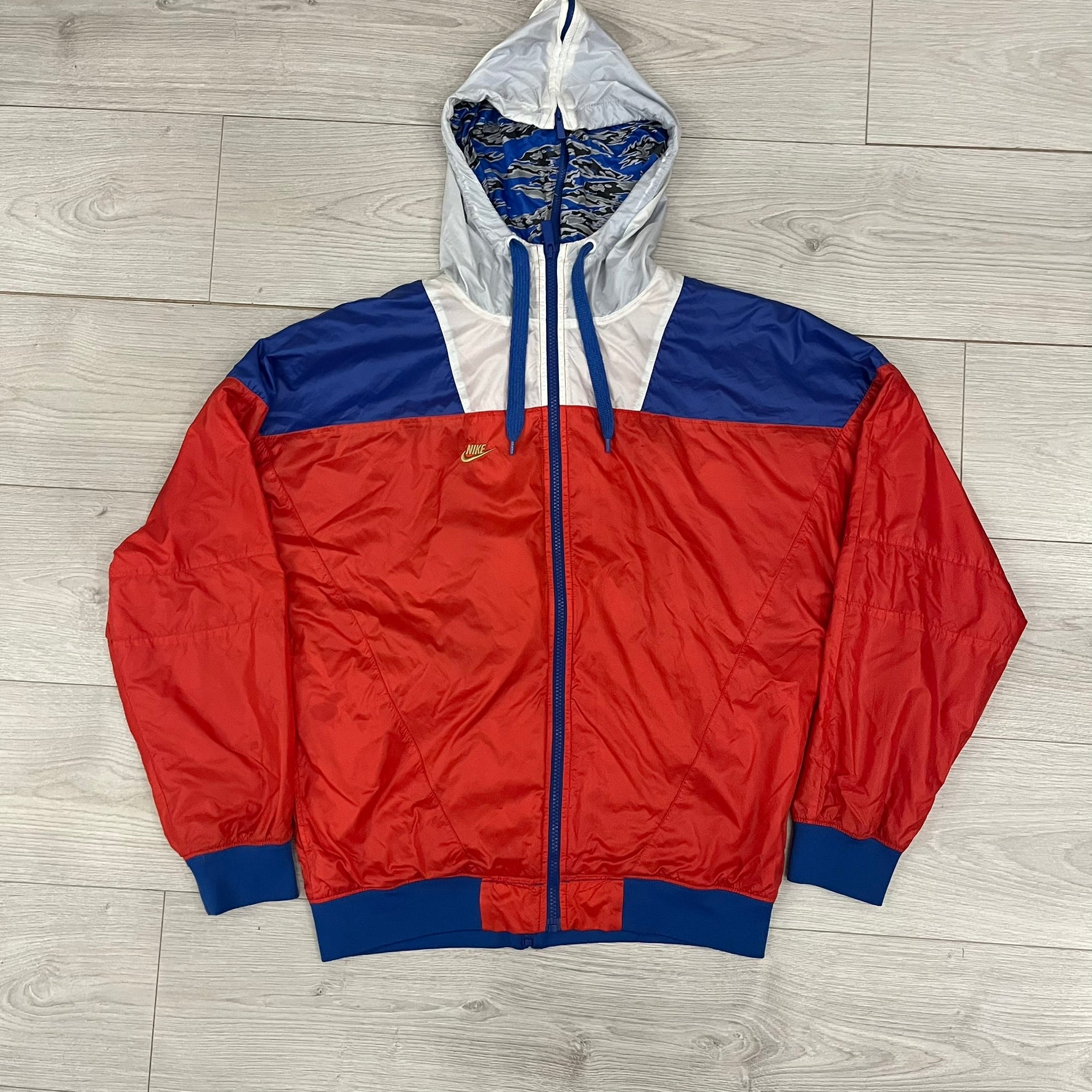 Nike Red/Blue/White Split Windbreaker featuring a modular design with front and back zippers that allow attachment to compatible jackets. The layout includes dominant red panels across the sleeves and upper front, contrasted with blue accents, a white hood, and a blue-white-black camo hood lining. Finished with a gold embroidered Nike logo on the chest.