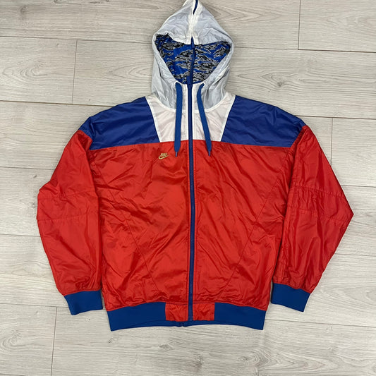 Nike Red/Blue/White Split Windbreaker featuring a modular design with front and back zippers that allow attachment to compatible jackets. The layout includes dominant red panels across the sleeves and upper front, contrasted with blue accents, a white hood, and a blue-white-black camo hood lining. Finished with a gold embroidered Nike logo on the chest.
