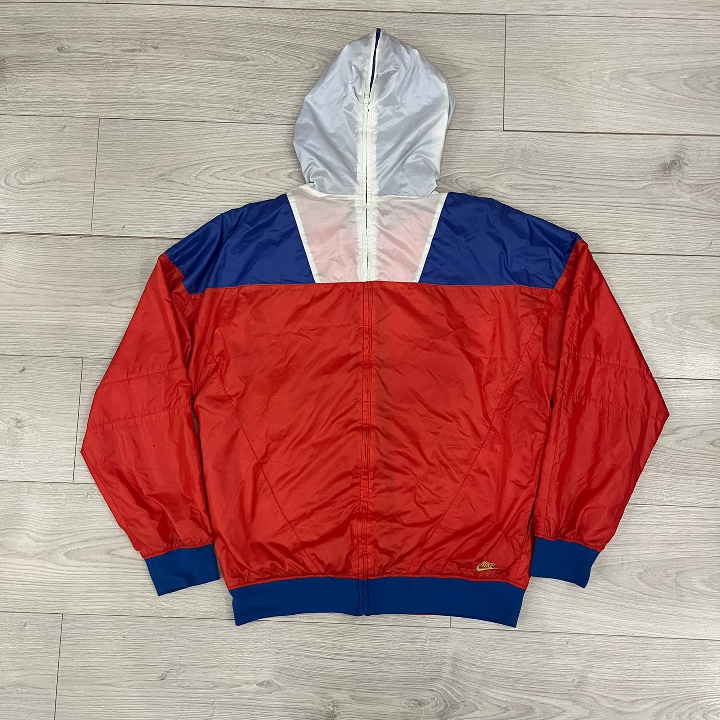 Nike Red/Blue/White Split Windbreaker featuring a modular design with front and back zippers that allow attachment to compatible jackets. The layout includes dominant red panels across the sleeves and upper front, contrasted with blue accents, a white hood, and a blue-white-black camo hood lining. Finished with a gold embroidered Nike logo on the chest.