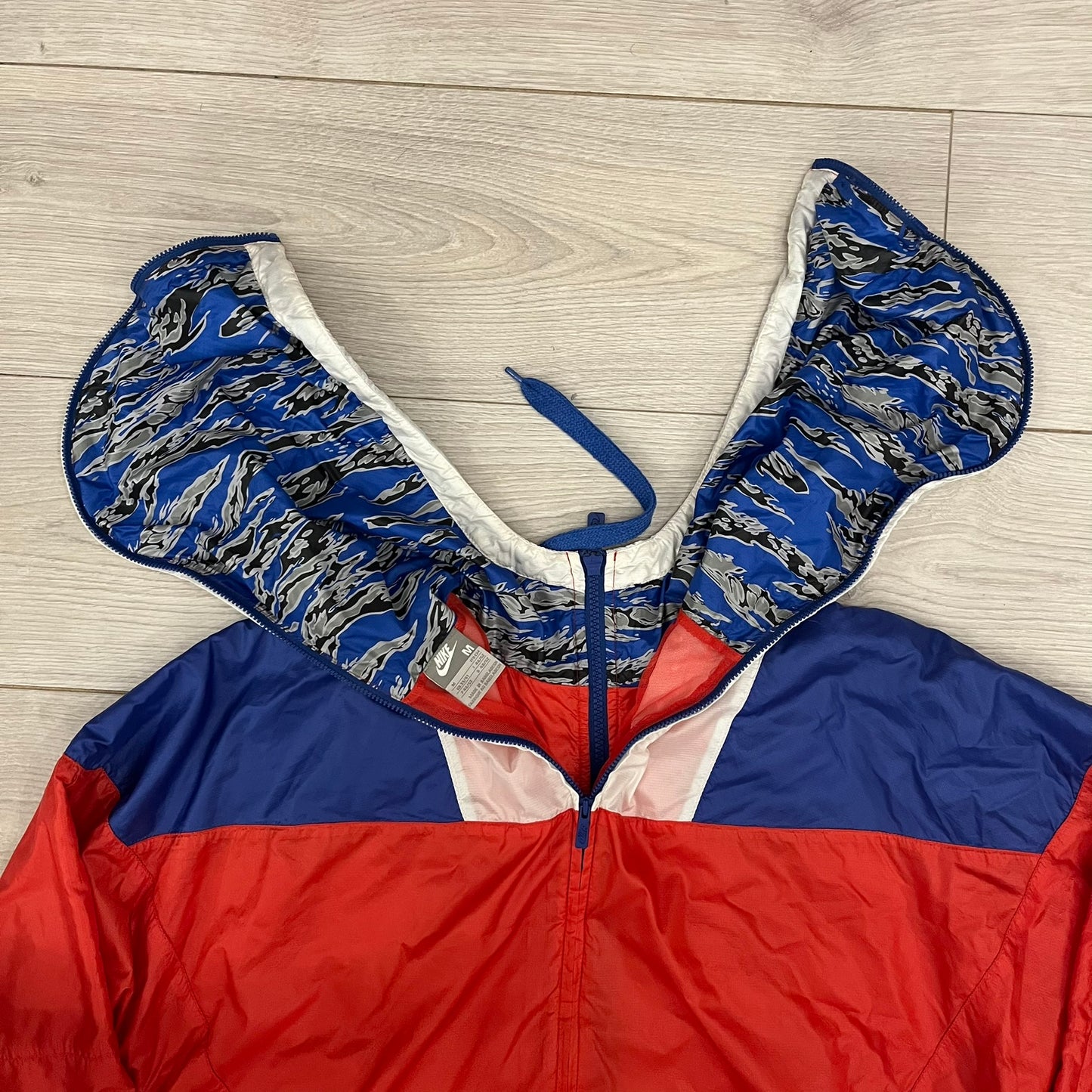 Nike Red/Blue/White Split Windbreaker featuring a modular design with front and back zippers that allow attachment to compatible jackets. The layout includes dominant red panels across the sleeves and upper front, contrasted with blue accents, a white hood, and a blue-white-black camo hood lining. Finished with a gold embroidered Nike logo on the chest.