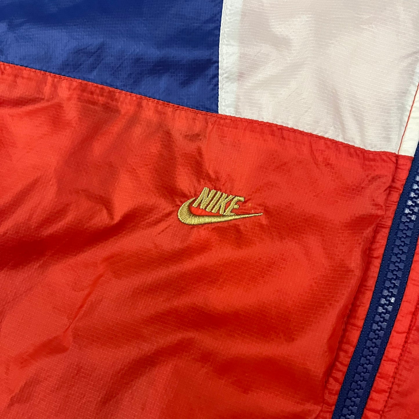 Nike Red/Blue/White Split Windbreaker featuring a modular design with front and back zippers that allow attachment to compatible jackets. The layout includes dominant red panels across the sleeves and upper front, contrasted with blue accents, a white hood, and a blue-white-black camo hood lining. Finished with a gold embroidered Nike logo on the chest.