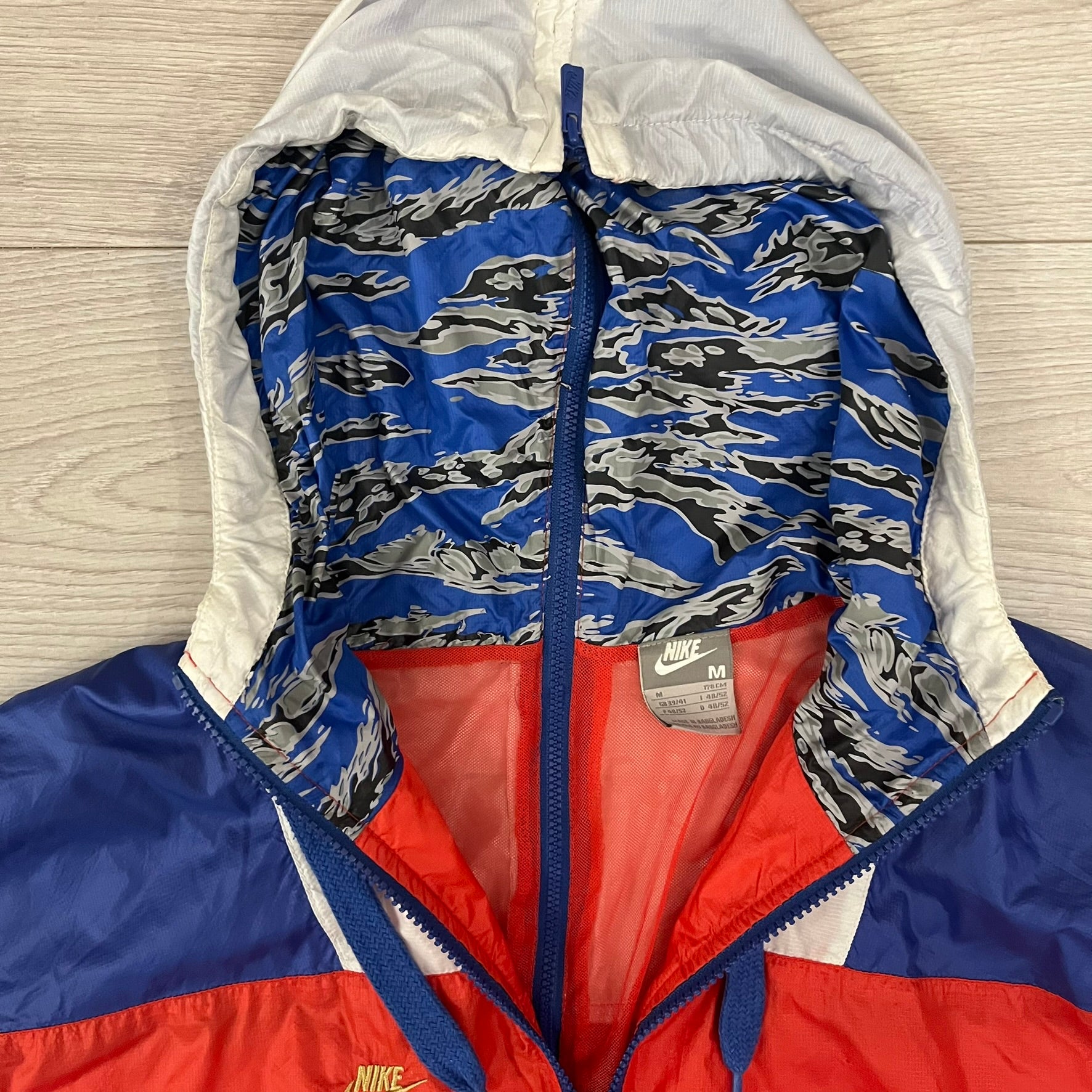 Nike Red/Blue/White Split Windbreaker featuring a modular design with front and back zippers that allow attachment to compatible jackets. The layout includes dominant red panels across the sleeves and upper front, contrasted with blue accents, a white hood, and a blue-white-black camo hood lining. Finished with a gold embroidered Nike logo on the chest.