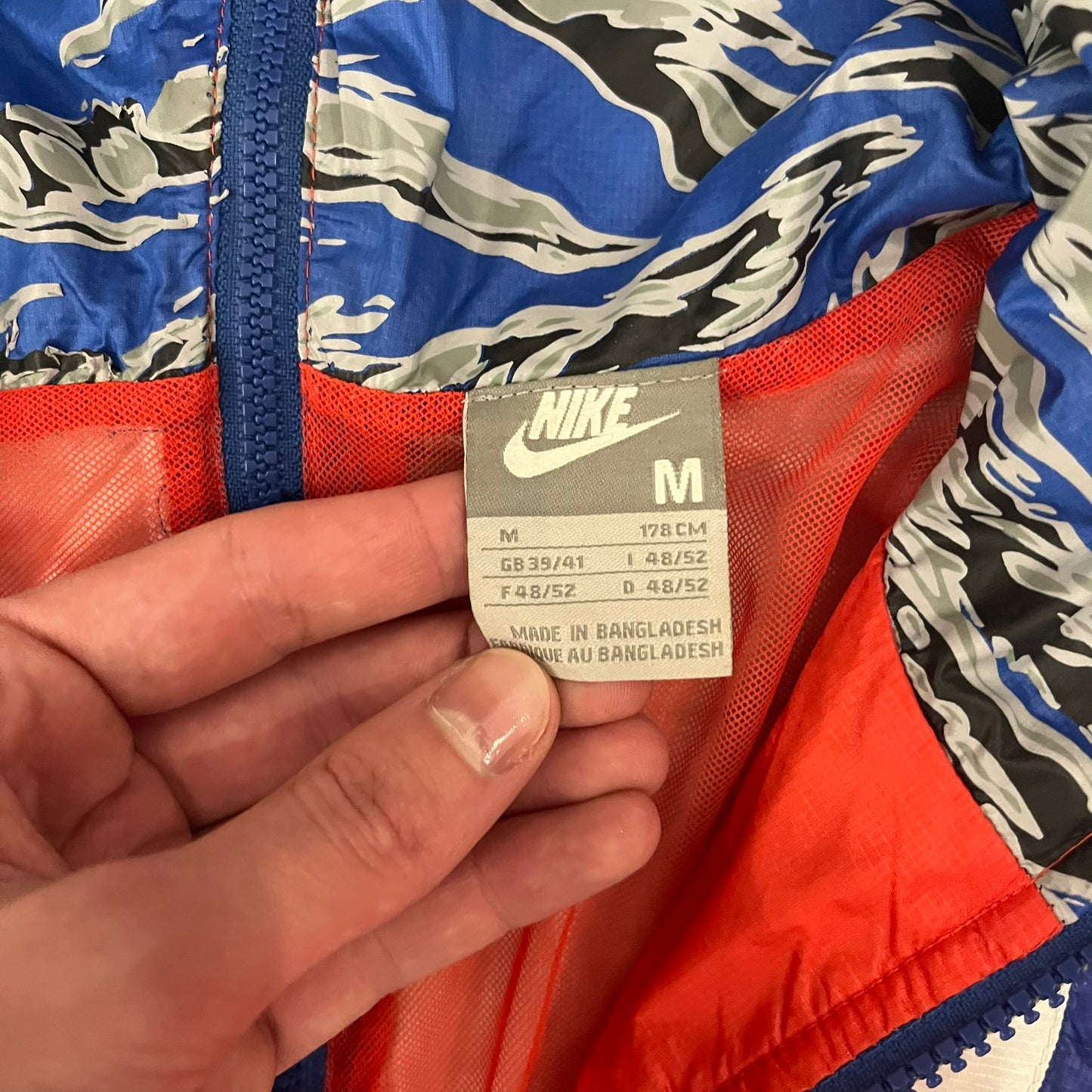 Nike Red/Blue/White Split Windbreaker featuring a modular design with front and back zippers that allow attachment to compatible jackets. The layout includes dominant red panels across the sleeves and upper front, contrasted with blue accents, a white hood, and a blue-white-black camo hood lining. Finished with a gold embroidered Nike logo on the chest.