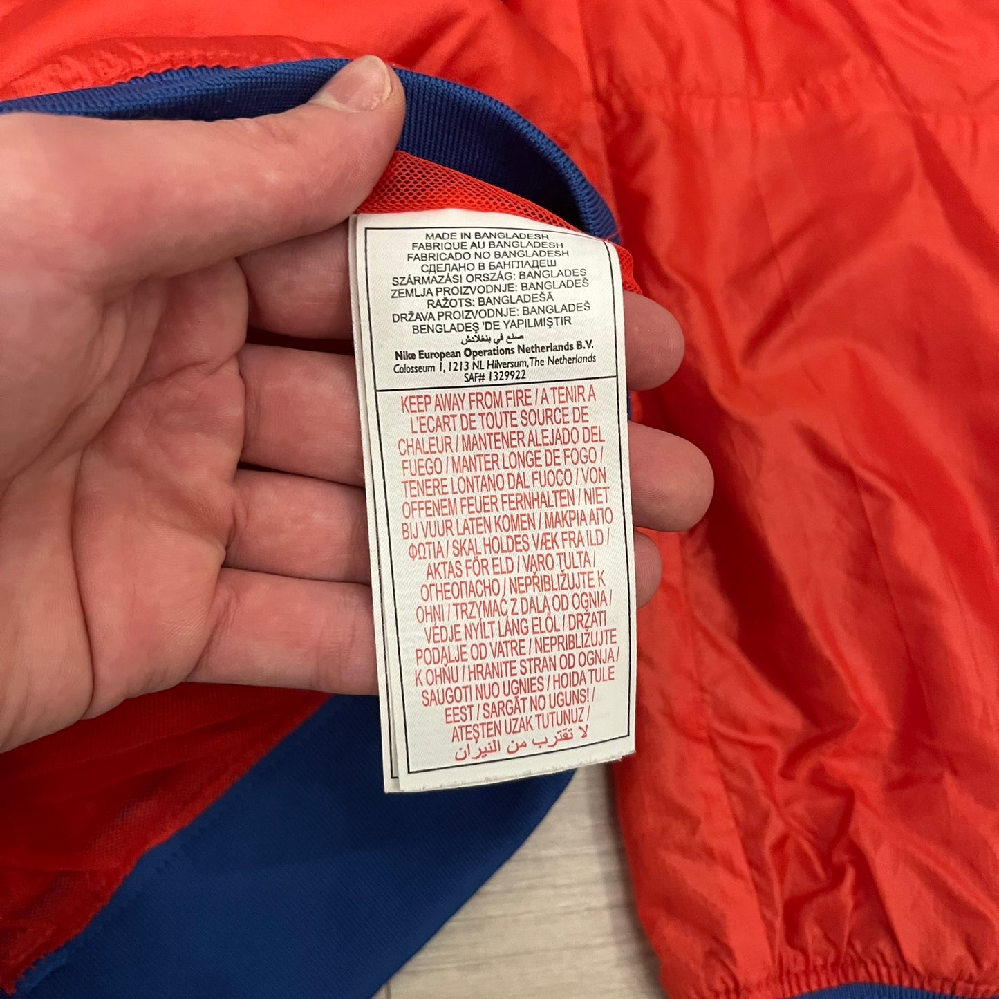 Nike Red/Blue/White Split Windbreaker featuring a modular design with front and back zippers that allow attachment to compatible jackets. The layout includes dominant red panels across the sleeves and upper front, contrasted with blue accents, a white hood, and a blue-white-black camo hood lining. Finished with a gold embroidered Nike logo on the chest.