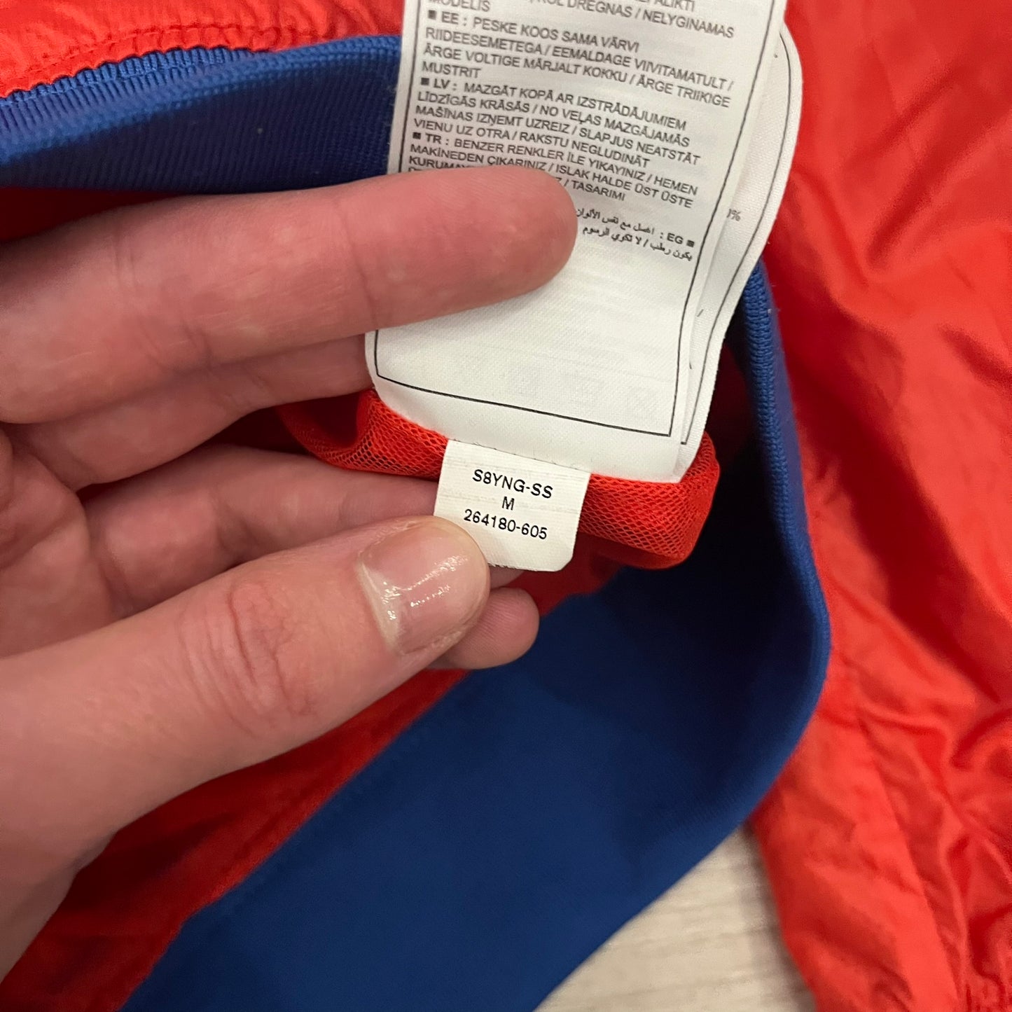 Nike Red/Blue/White Split Windbreaker featuring a modular design with front and back zippers that allow attachment to compatible jackets. The layout includes dominant red panels across the sleeves and upper front, contrasted with blue accents, a white hood, and a blue-white-black camo hood lining. Finished with a gold embroidered Nike logo on the chest.