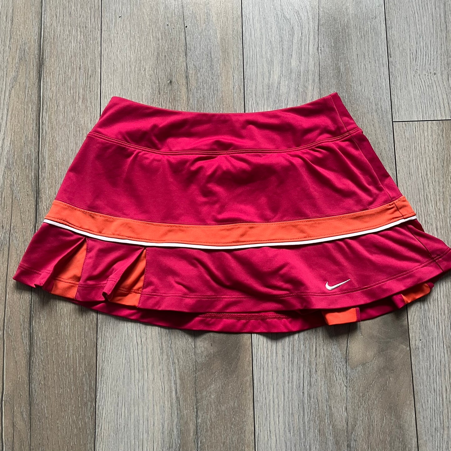 Nike Smash Classic Pink/Orange Tennis Skirt is a women’s tennis skort with built-in inner shorts. The design is constructed in three sections: a dark pink waistband and upper panel, an orange middle stripe, and a lower dark pink layer with two cutouts revealing orange fabric underneath. A white Nike Swoosh is placed at the lower hem, with a small Nike Court logo on the side.