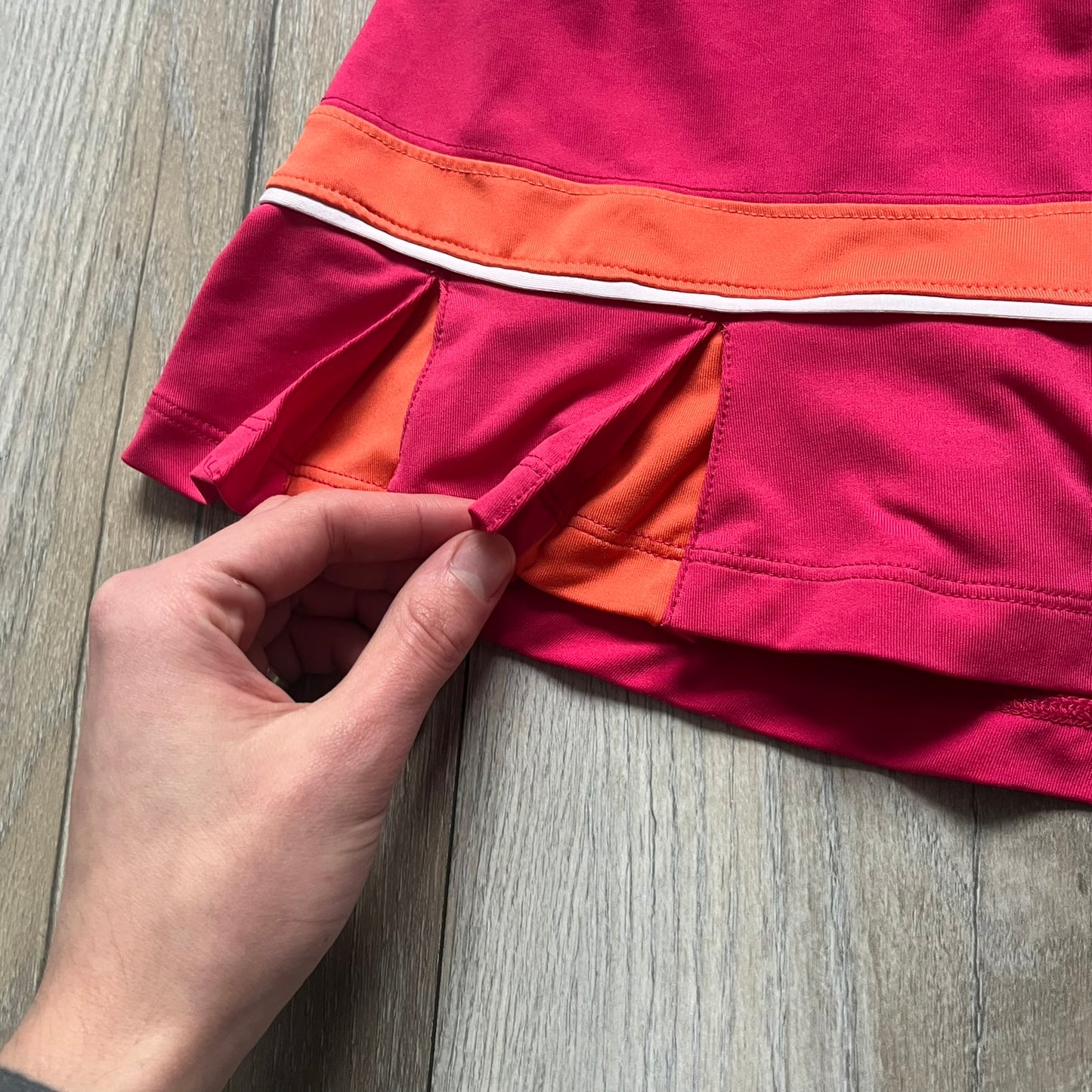 Nike Smash Classic Pink/Orange Tennis Skirt is a women’s tennis skort with built-in inner shorts. The design is constructed in three sections: a dark pink waistband and upper panel, an orange middle stripe, and a lower dark pink layer with two cutouts revealing orange fabric underneath. A white Nike Swoosh is placed at the lower hem, with a small Nike Court logo on the side.