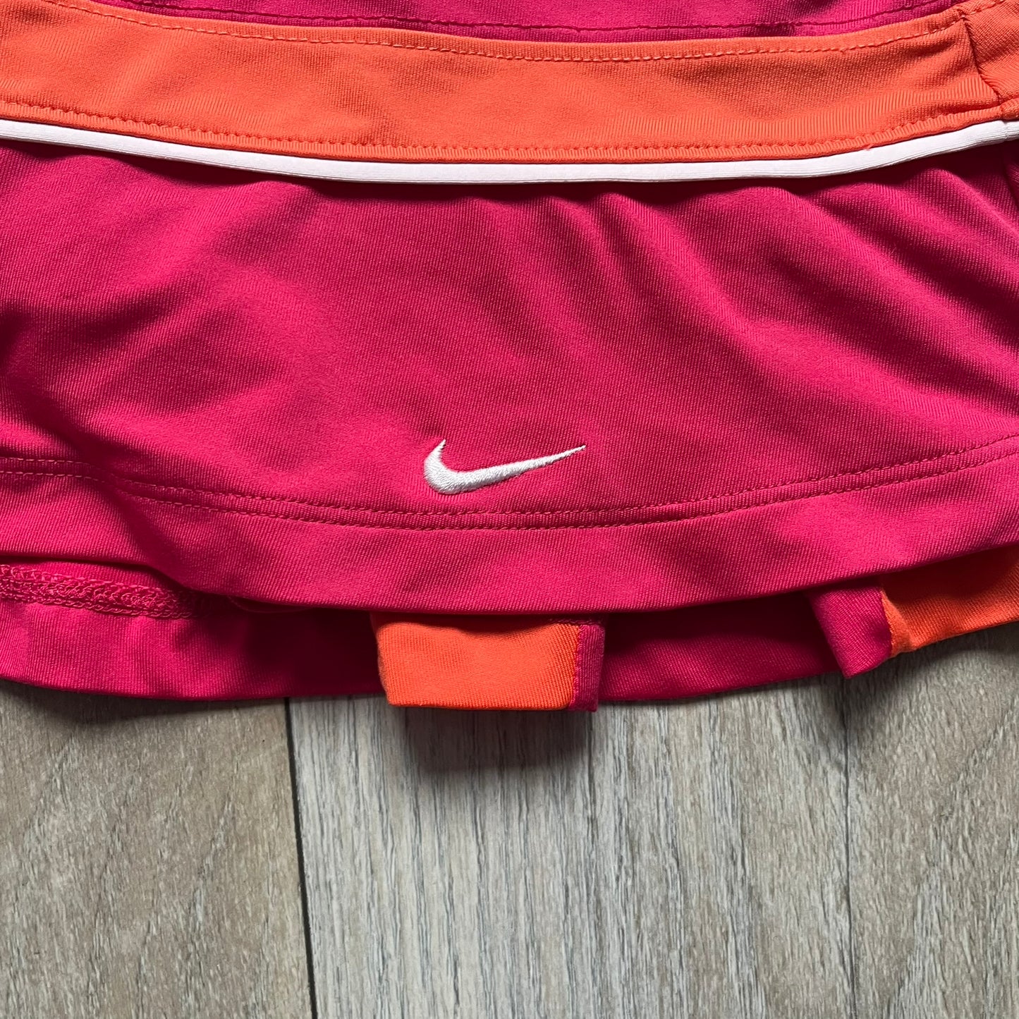 Nike Smash Classic Pink/Orange Tennis Skirt is a women’s tennis skort with built-in inner shorts. The design is constructed in three sections: a dark pink waistband and upper panel, an orange middle stripe, and a lower dark pink layer with two cutouts revealing orange fabric underneath. A white Nike Swoosh is placed at the lower hem, with a small Nike Court logo on the side.