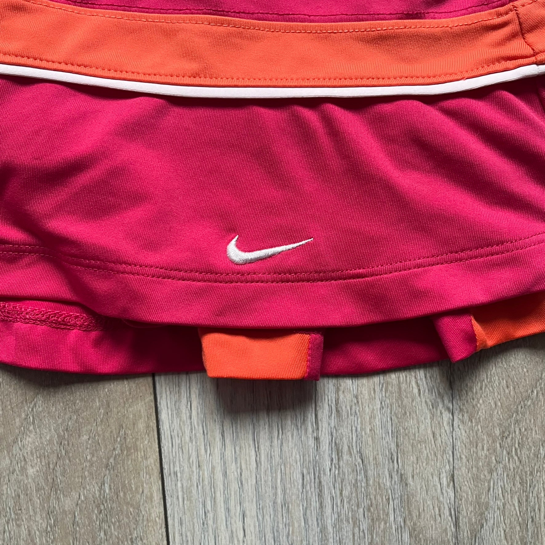 Nike Smash Classic Pink/Orange Tennis Skirt is a women’s tennis skort with built-in inner shorts. The design is constructed in three sections: a dark pink waistband and upper panel, an orange middle stripe, and a lower dark pink layer with two cutouts revealing orange fabric underneath. A white Nike Swoosh is placed at the lower hem, with a small Nike Court logo on the side.