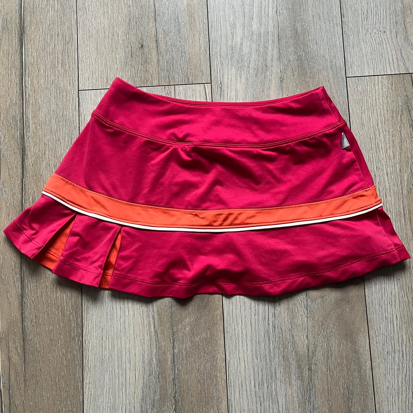 Nike Smash Classic Pink/Orange Tennis Skirt is a women’s tennis skort with built-in inner shorts. The design is constructed in three sections: a dark pink waistband and upper panel, an orange middle stripe, and a lower dark pink layer with two cutouts revealing orange fabric underneath. A white Nike Swoosh is placed at the lower hem, with a small Nike Court logo on the side.