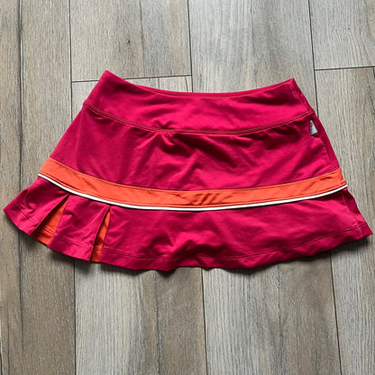 Nike Smash Classic Pink/Orange Tennis Skirt is a women’s tennis skort with built-in inner shorts. The design is constructed in three sections: a dark pink waistband and upper panel, an orange middle stripe, and a lower dark pink layer with two cutouts revealing orange fabric underneath. A white Nike Swoosh is placed at the lower hem, with a small Nike Court logo on the side.