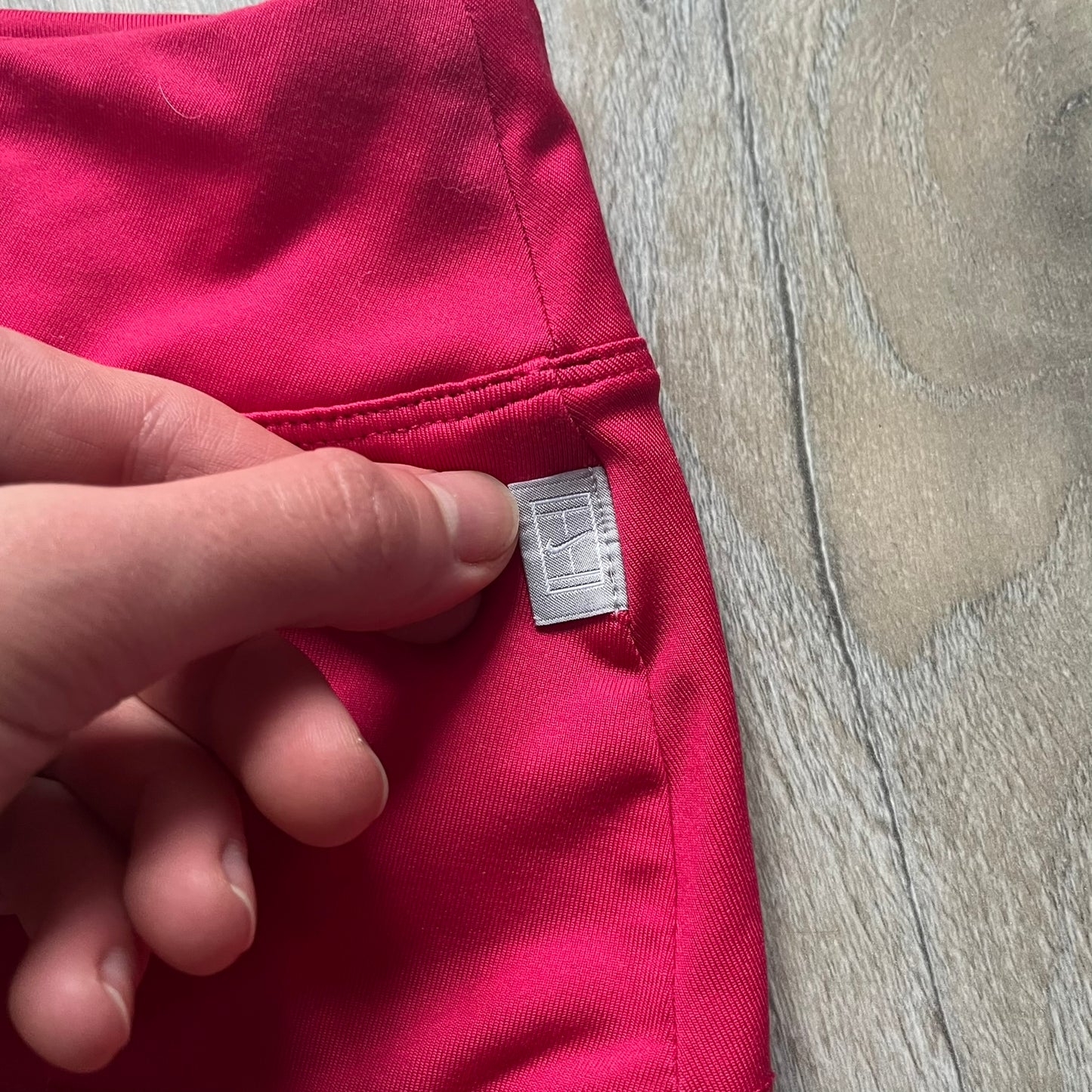 Nike Smash Classic Pink/Orange Tennis Skirt is a women’s tennis skort with built-in inner shorts. The design is constructed in three sections: a dark pink waistband and upper panel, an orange middle stripe, and a lower dark pink layer with two cutouts revealing orange fabric underneath. A white Nike Swoosh is placed at the lower hem, with a small Nike Court logo on the side.