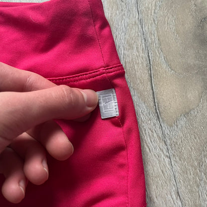 Nike Smash Classic Pink/Orange Tennis Skirt is a women’s tennis skort with built-in inner shorts. The design is constructed in three sections: a dark pink waistband and upper panel, an orange middle stripe, and a lower dark pink layer with two cutouts revealing orange fabric underneath. A white Nike Swoosh is placed at the lower hem, with a small Nike Court logo on the side.