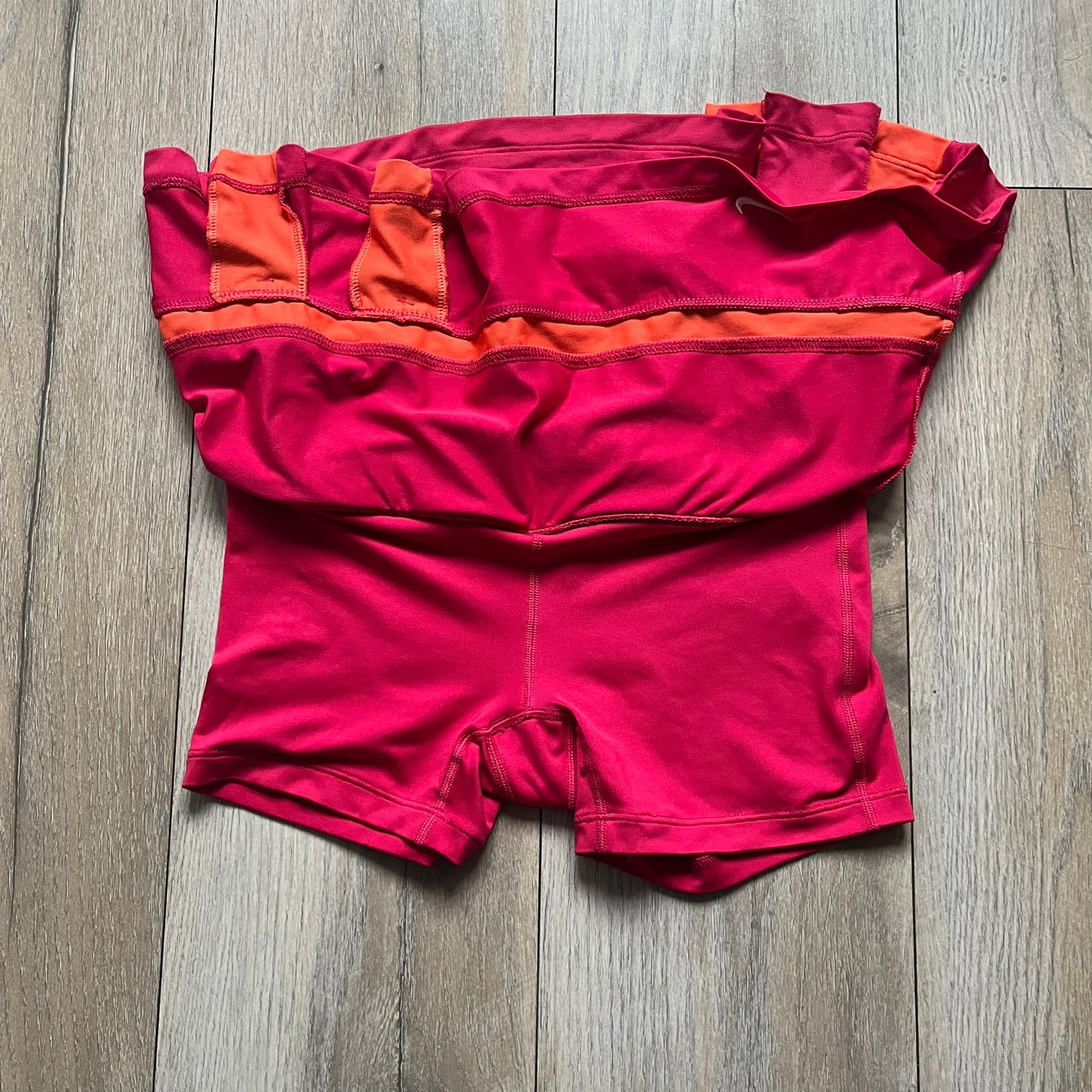 Nike Smash Classic Pink/Orange Tennis Skirt is a women’s tennis skort with built-in inner shorts. The design is constructed in three sections: a dark pink waistband and upper panel, an orange middle stripe, and a lower dark pink layer with two cutouts revealing orange fabric underneath. A white Nike Swoosh is placed at the lower hem, with a small Nike Court logo on the side.