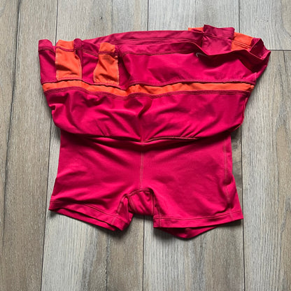 Nike Smash Classic Pink/Orange Tennis Skirt is a women’s tennis skort with built-in inner shorts. The design is constructed in three sections: a dark pink waistband and upper panel, an orange middle stripe, and a lower dark pink layer with two cutouts revealing orange fabric underneath. A white Nike Swoosh is placed at the lower hem, with a small Nike Court logo on the side.