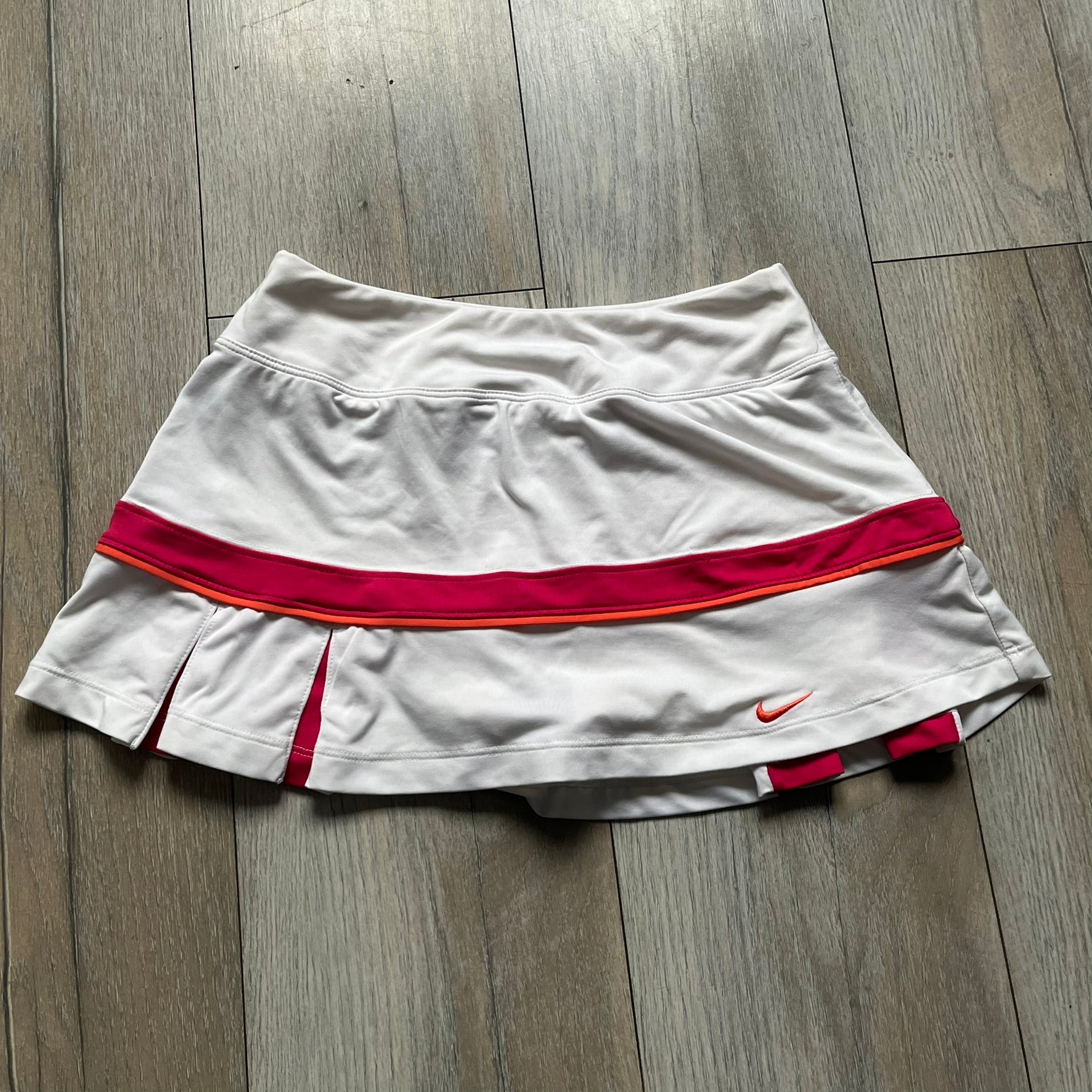 Nike Smash Classic White/Red Tennis Skirt is a women’s tennis skort with built-in inner shorts. The design is constructed in three sections: a white waistband and upper panel, a red middle stripe, and a lower white layer with two cutouts revealing red fabric underneath. A white Nike Swoosh is placed at the lower hem, with a small Nike Court logo on the side.