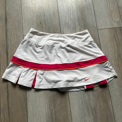 Nike Smash Classic White/Red Tennis Skirt is a women’s tennis skort with built-in inner shorts. The design is constructed in three sections: a white waistband and upper panel, a red middle stripe, and a lower white layer with two cutouts revealing red fabric underneath. A white Nike Swoosh is placed at the lower hem, with a small Nike Court logo on the side.
