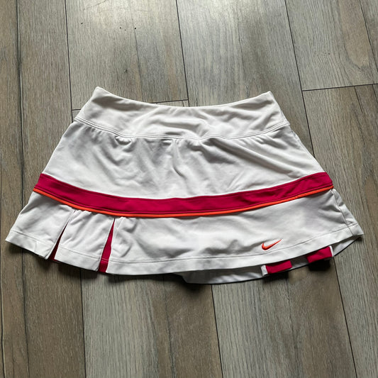 Nike Smash Classic White/Red Tennis Skirt is a women’s tennis skort with built-in inner shorts. The design is constructed in three sections: a white waistband and upper panel, a red middle stripe, and a lower white layer with two cutouts revealing red fabric underneath. A white Nike Swoosh is placed at the lower hem, with a small Nike Court logo on the side.