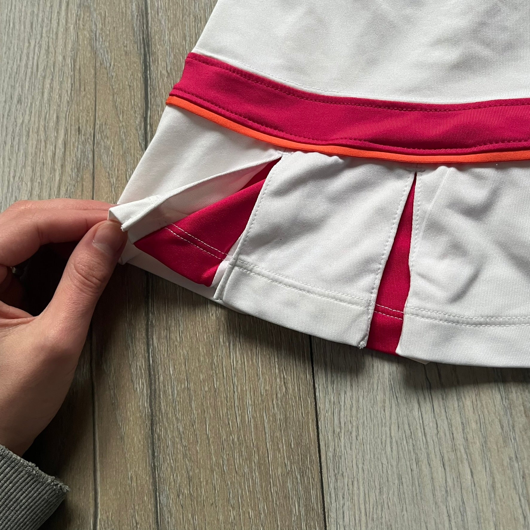 Nike Smash Classic White/Red Tennis Skirt is a women’s tennis skort with built-in inner shorts. The design is constructed in three sections: a white waistband and upper panel, a red middle stripe, and a lower white layer with two cutouts revealing red fabric underneath. A white Nike Swoosh is placed at the lower hem, with a small Nike Court logo on the side.