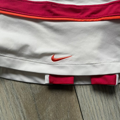 Nike Smash Classic White/Red Tennis Skirt is a women’s tennis skort with built-in inner shorts. The design is constructed in three sections: a white waistband and upper panel, a red middle stripe, and a lower white layer with two cutouts revealing red fabric underneath. A white Nike Swoosh is placed at the lower hem, with a small Nike Court logo on the side.