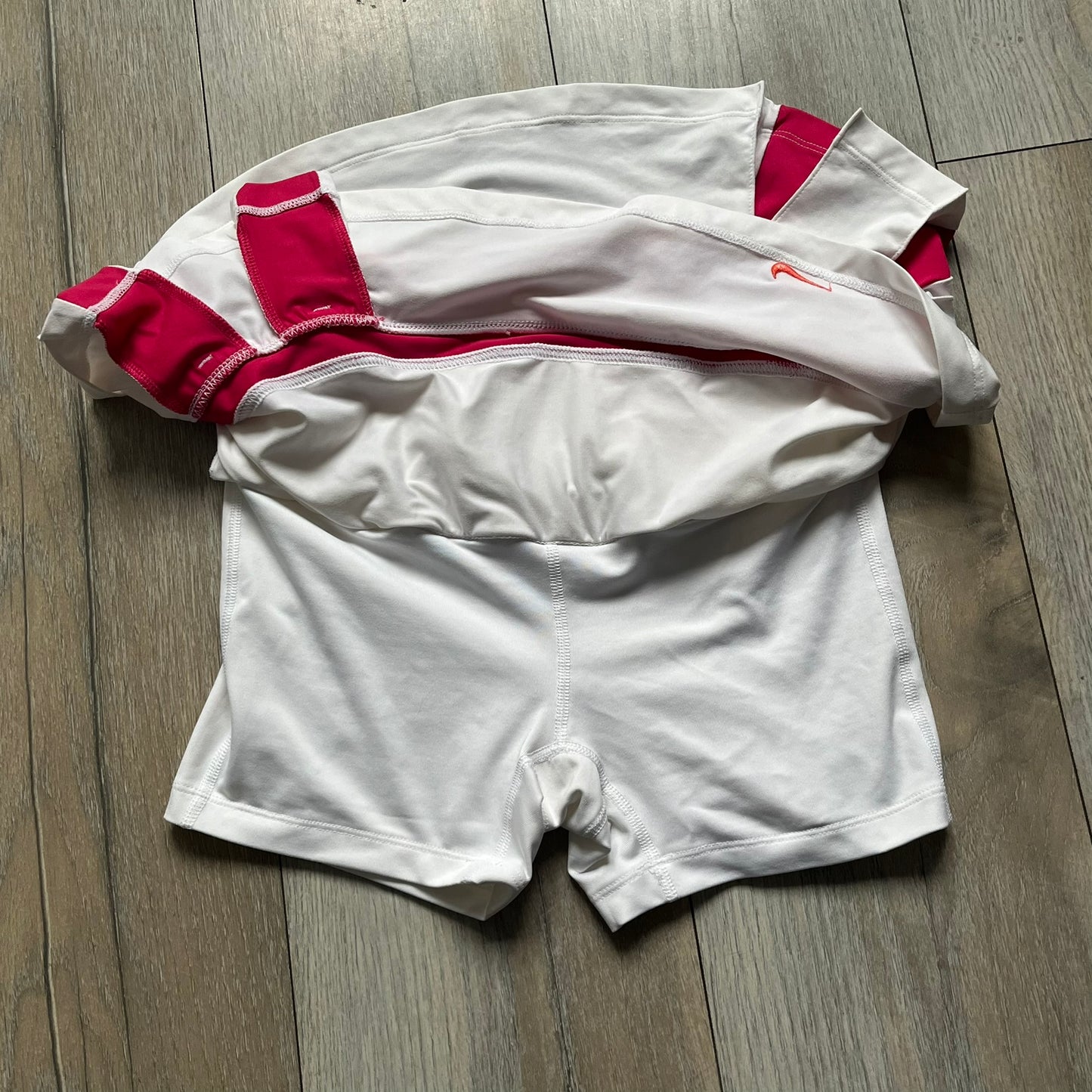 Nike Smash Classic White/Red Tennis Skirt is a women’s tennis skort with built-in inner shorts. The design is constructed in three sections: a white waistband and upper panel, a red middle stripe, and a lower white layer with two cutouts revealing red fabric underneath. A white Nike Swoosh is placed at the lower hem, with a small Nike Court logo on the side.