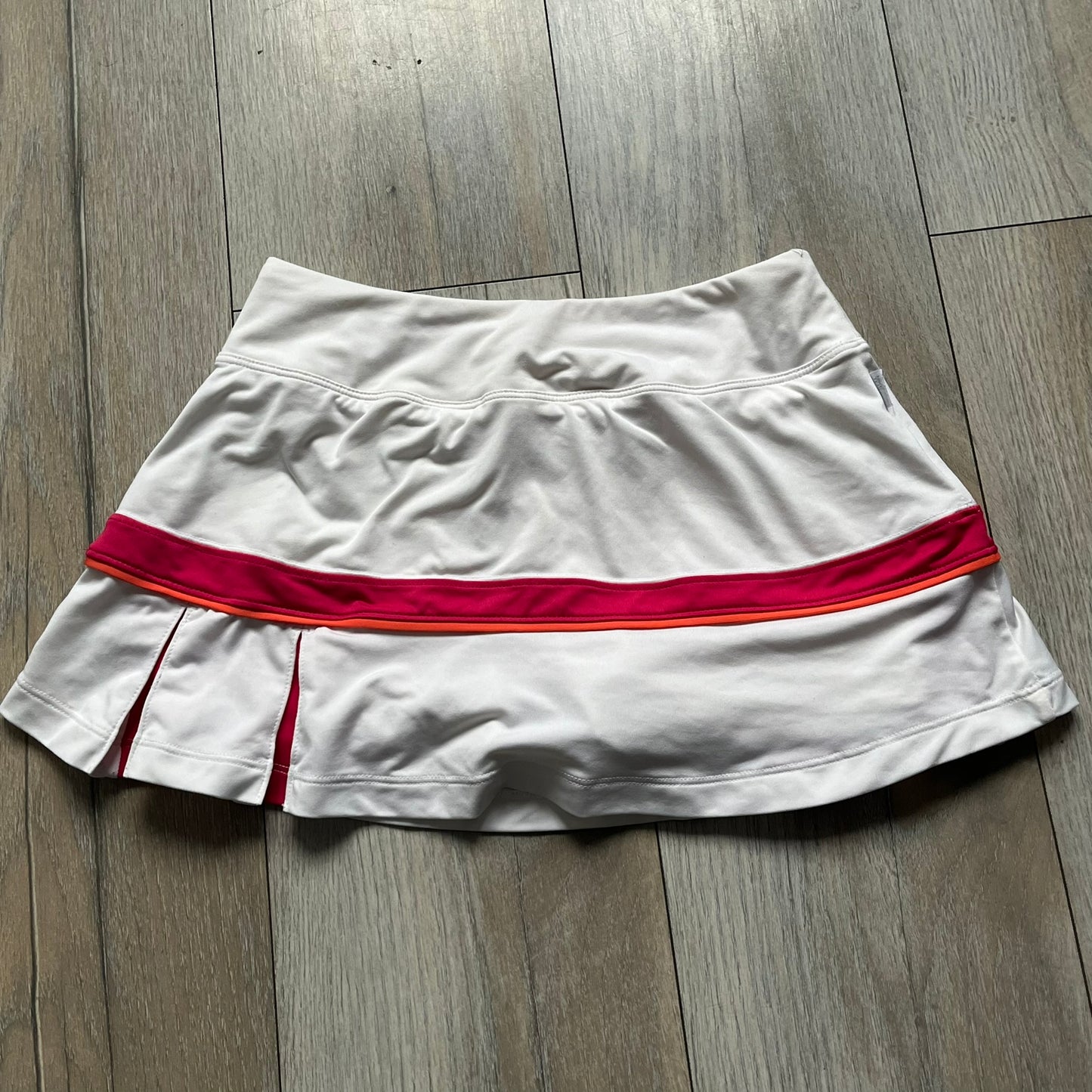 Nike Smash Classic White/Red Tennis Skirt is a women’s tennis skort with built-in inner shorts. The design is constructed in three sections: a white waistband and upper panel, a red middle stripe, and a lower white layer with two cutouts revealing red fabric underneath. A white Nike Swoosh is placed at the lower hem, with a small Nike Court logo on the side.