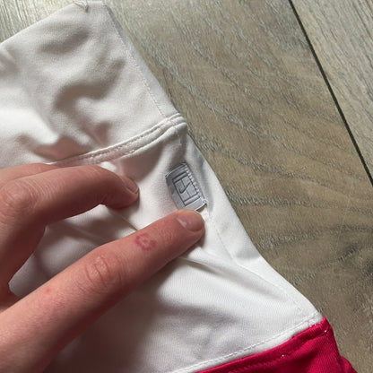 Nike Smash Classic White/Red Tennis Skirt is a women’s tennis skort with built-in inner shorts. The design is constructed in three sections: a white waistband and upper panel, a red middle stripe, and a lower white layer with two cutouts revealing red fabric underneath. A white Nike Swoosh is placed at the lower hem, with a small Nike Court logo on the side.