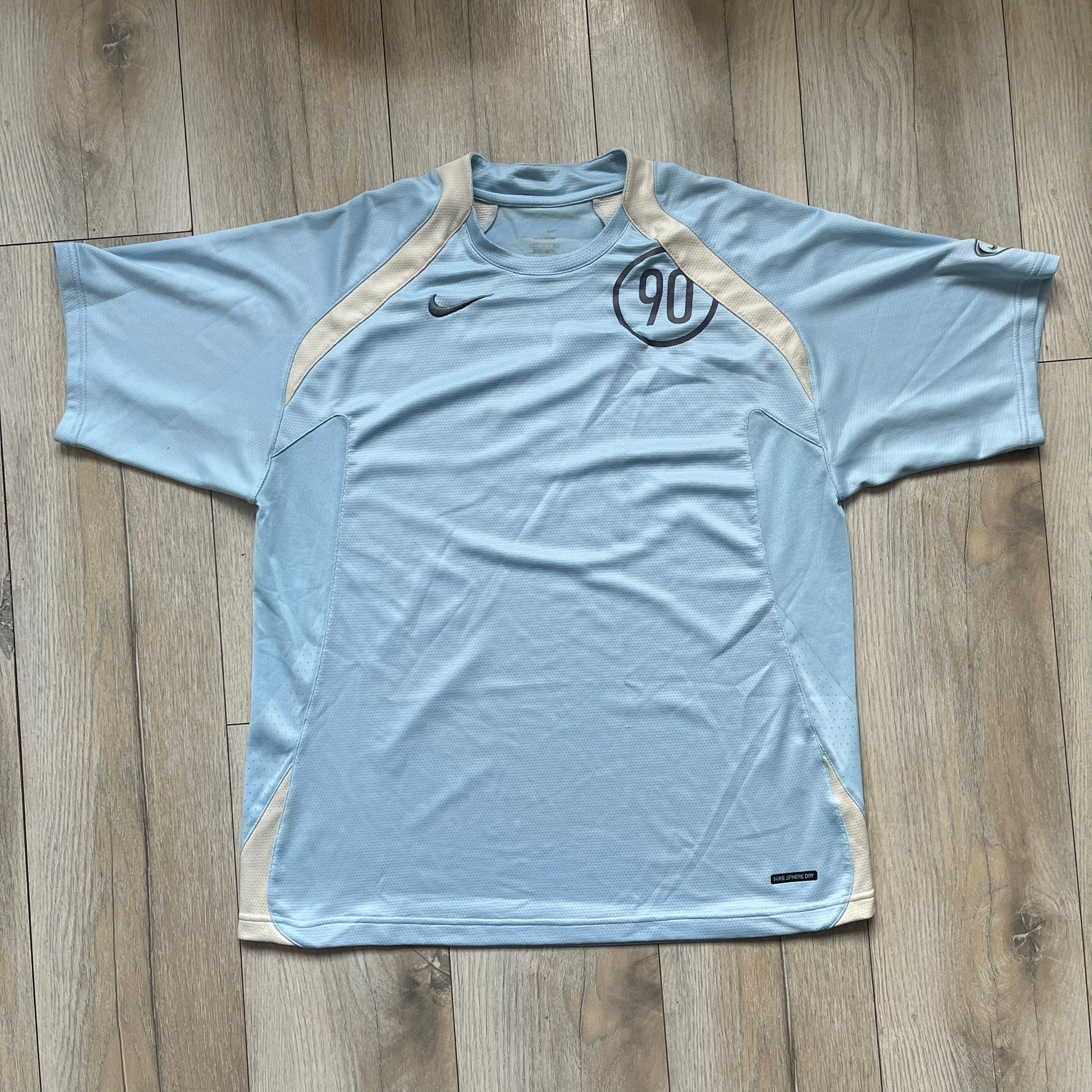 Nike Sphere Dry 90’s Baby Blue Tee is a baby blue T-shirt featuring gray and white stripe detailing. The front includes a dark gray Nike logo alongside a “90” graphic, with an additional Nike logo on the sleeve. The back displays a large light blue “90” print