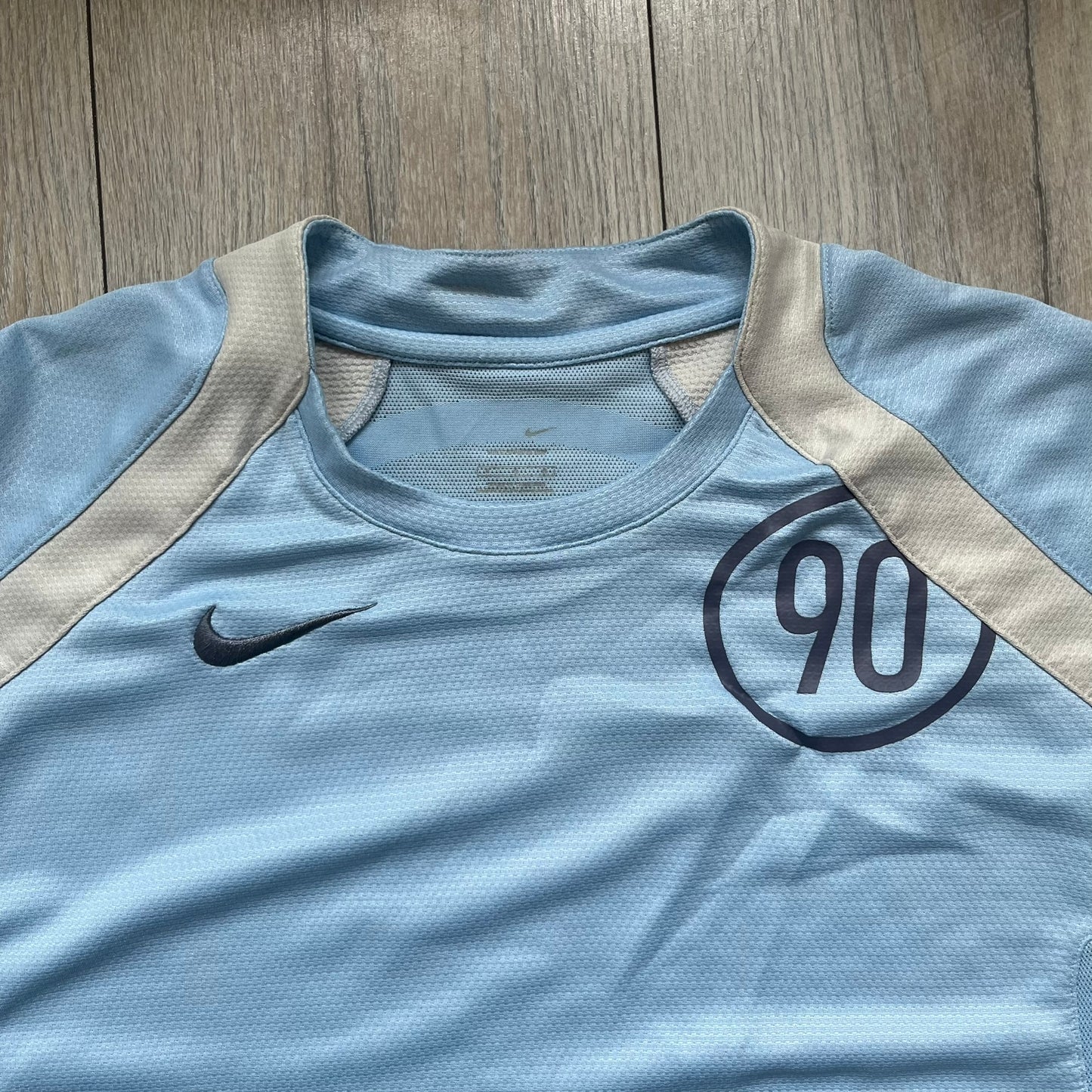 Nike Sphere Dry 90’s Baby Blue Tee is a baby blue T-shirt featuring gray and white stripe detailing. The front includes a dark gray Nike logo alongside a “90” graphic, with an additional Nike logo on the sleeve. The back displays a large light blue “90” print