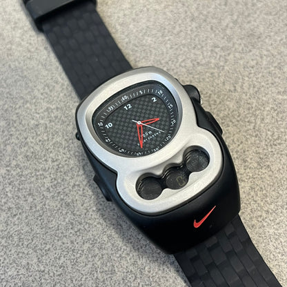 Nike Steelhead 2000’s Sport Chrono VG02-5000 Watch has a gray and black checkered analog dial with red hands, set in a rounded triangle shape. Below it is a small digital screen made of three connected circles. The case is black and silver with a red Nike Swoosh at the bottom, and the original straps have been replaced with basic non-Nike rubber ones.