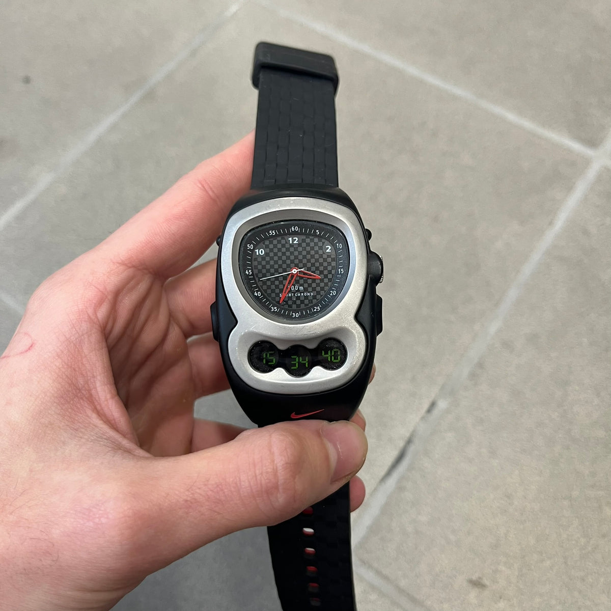 Nike Steelhead 2000’s Sport Chrono VG02-5000 Watch has a gray and black checkered analog dial with red hands, set in a rounded triangle shape. Below it is a small digital screen made of three connected circles. The case is black and silver with a red Nike Swoosh at the bottom, and the original straps have been replaced with basic non-Nike rubber ones.