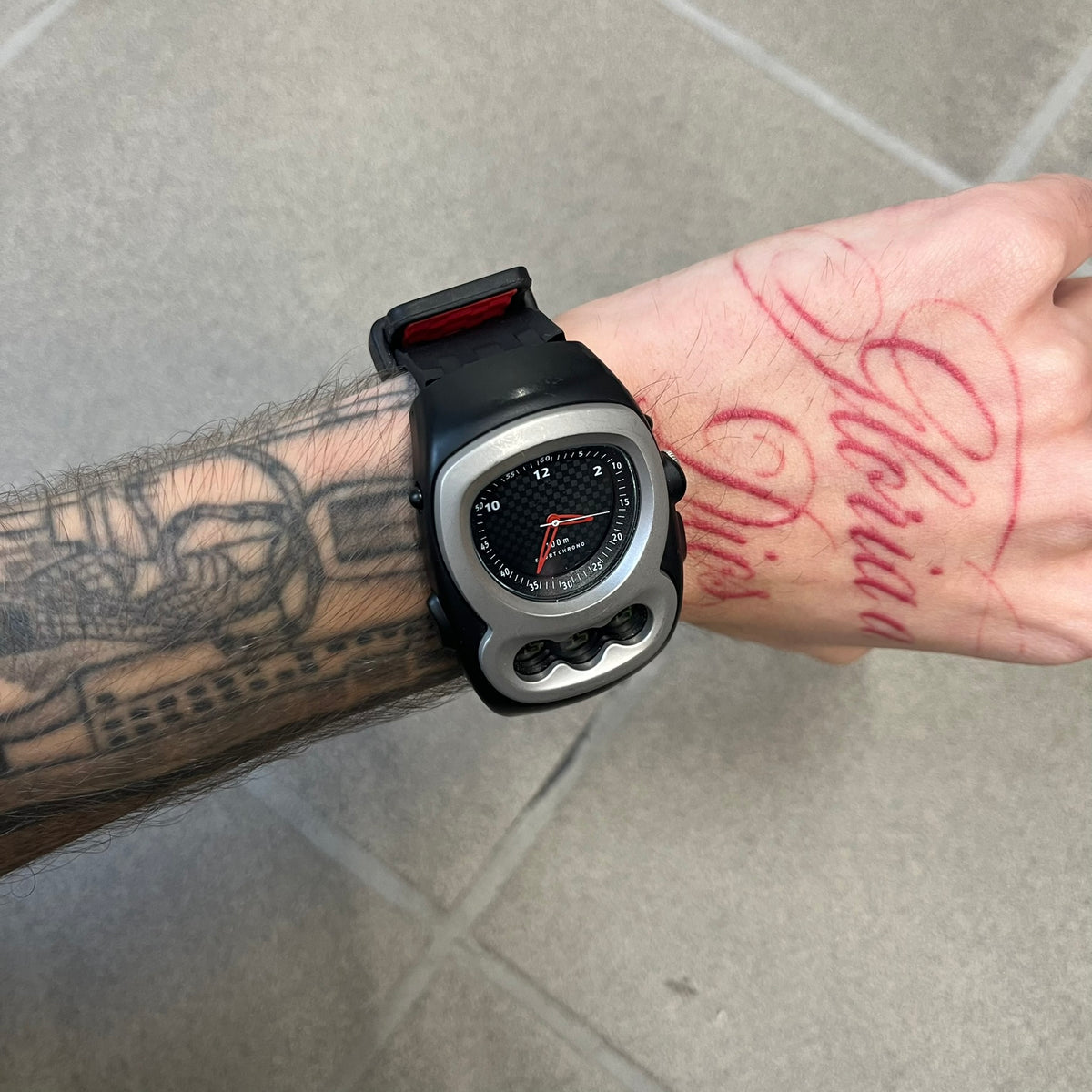 Nike Steelhead 2000’s Sport Chrono VG02-5000 Watch has a gray and black checkered analog dial with red hands, set in a rounded triangle shape. Below it is a small digital screen made of three connected circles. The case is black and silver with a red Nike Swoosh at the bottom, and the original straps have been replaced with basic non-Nike rubber ones.