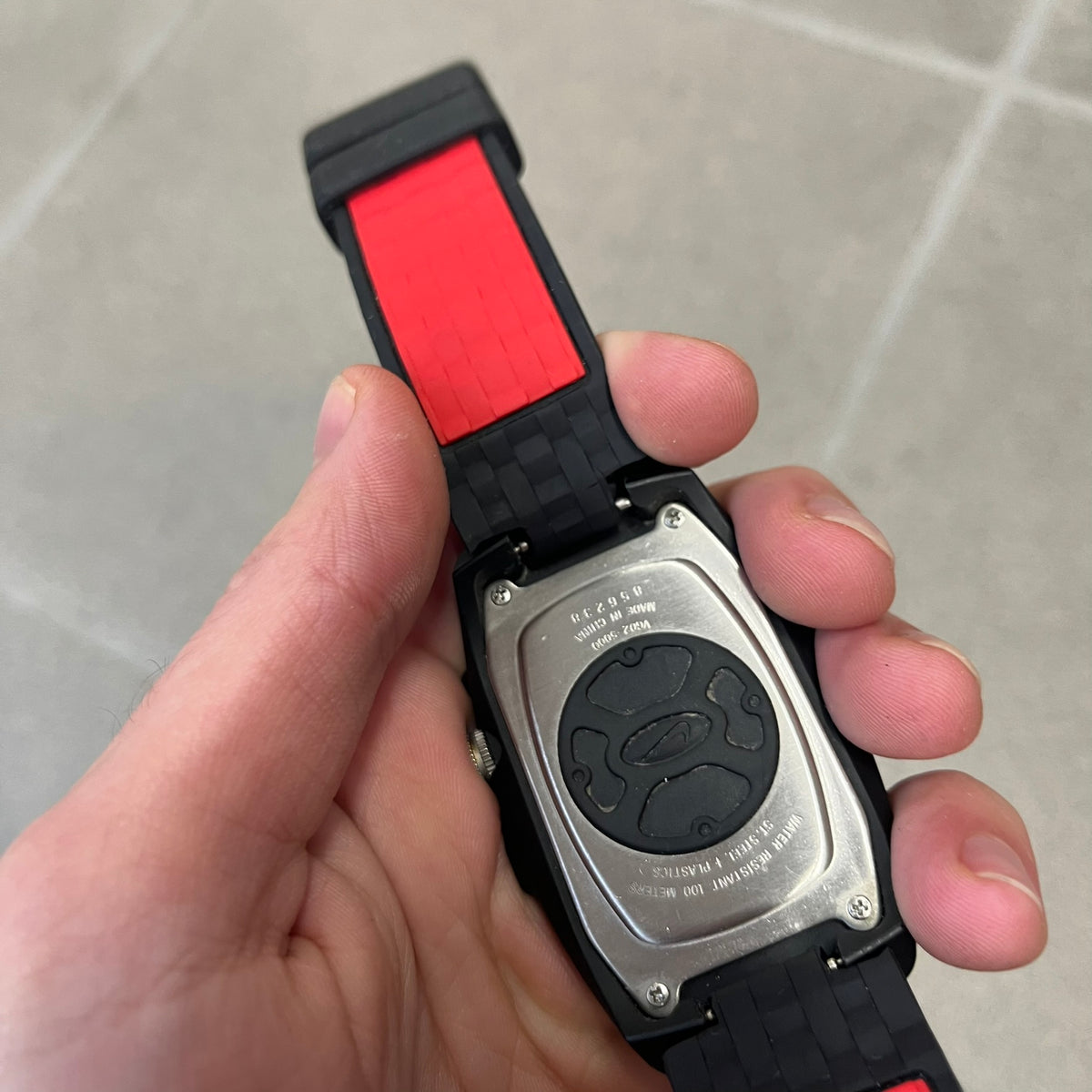 Nike Steelhead 2000’s Sport Chrono VG02-5000 Watch has a gray and black checkered analog dial with red hands, set in a rounded triangle shape. Below it is a small digital screen made of three connected circles. The case is black and silver with a red Nike Swoosh at the bottom, and the original straps have been replaced with basic non-Nike rubber ones.