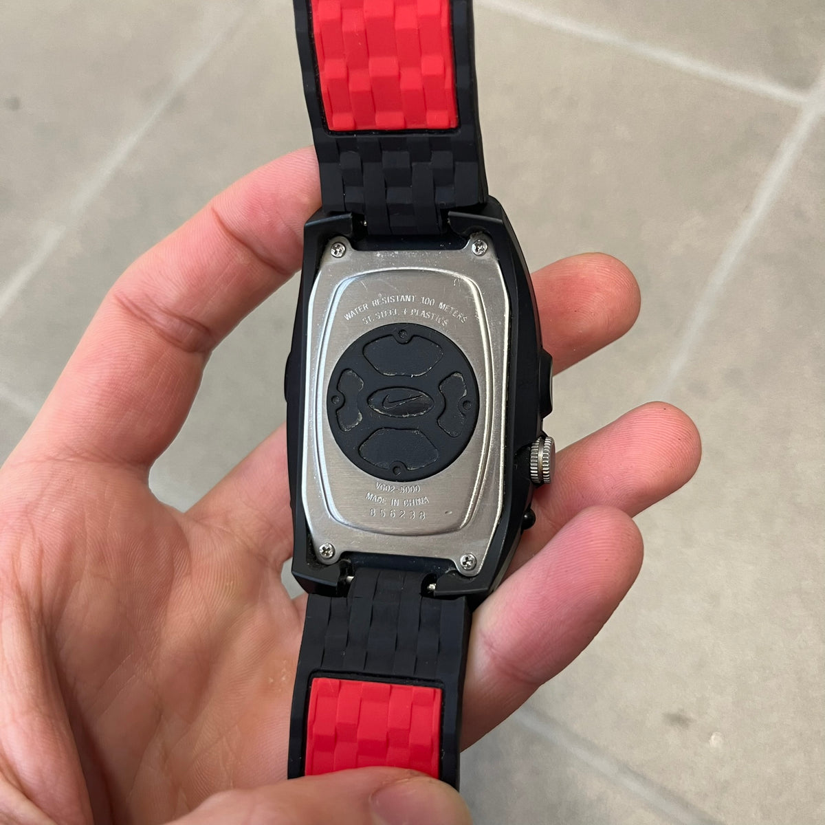 Nike Steelhead 2000’s Sport Chrono VG02-5000 Watch has a gray and black checkered analog dial with red hands, set in a rounded triangle shape. Below it is a small digital screen made of three connected circles. The case is black and silver with a red Nike Swoosh at the bottom, and the original straps have been replaced with basic non-Nike rubber ones.