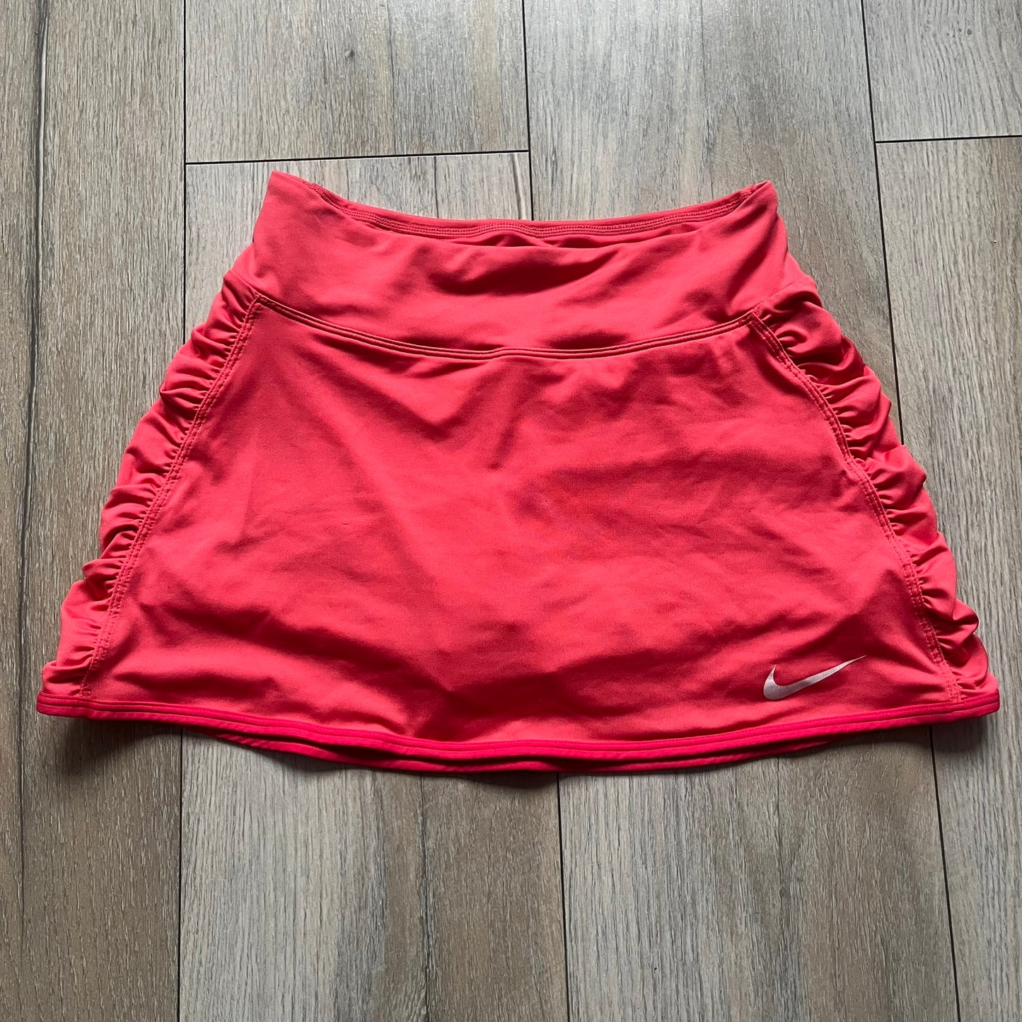 Nike Tie Break Knit 2012 Coral Tennis Skirt is a women’s tennis skort in coral featuring an integrated inner shorts layer. The skirt has a flared, creased design at the sides and a white Nike Swoosh at the lower hem. Designed for sporty performance, it combines lightweight knit construction with Nike’s 2012 Tie Break tennis line styling.