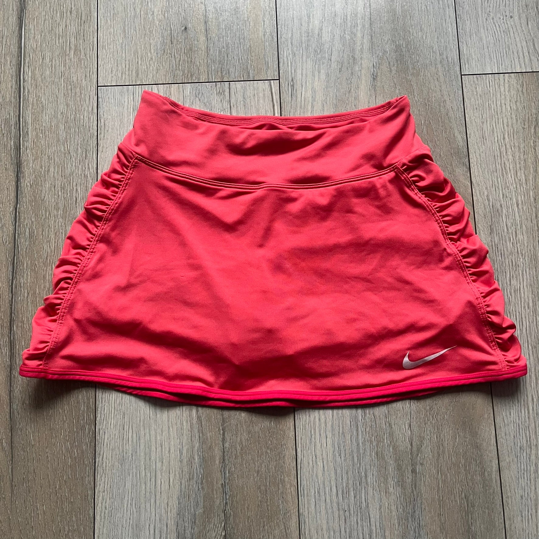 Nike Tie Break Knit 2012 Coral Tennis Skirt is a women’s tennis skort in coral featuring an integrated inner shorts layer. The skirt has a flared, creased design at the sides and a white Nike Swoosh at the lower hem. Designed for sporty performance, it combines lightweight knit construction with Nike’s 2012 Tie Break tennis line styling.