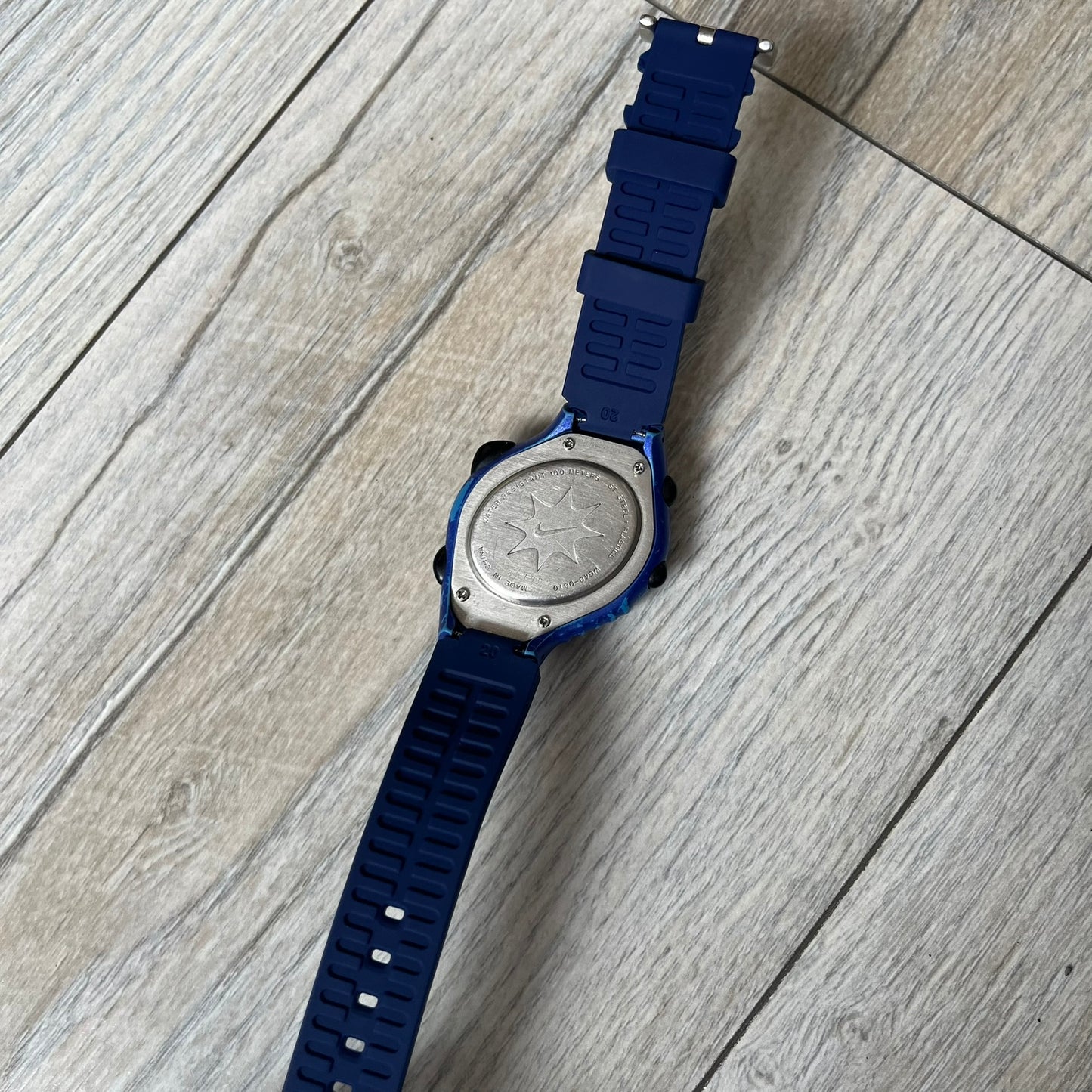 Nike Triax 120 WG40-0010 Digital Watch features a blue case with a black Nike Swoosh above the display and a black control button below it. The watch has a black digital screen with light-colored numerals and is fitted with navy-blue replacement straps that are not original Nike straps. Constructed from stainless steel and plastic materials, it offers 100-meter water resistance, 120-lap memory, and multiple timing and training modes.