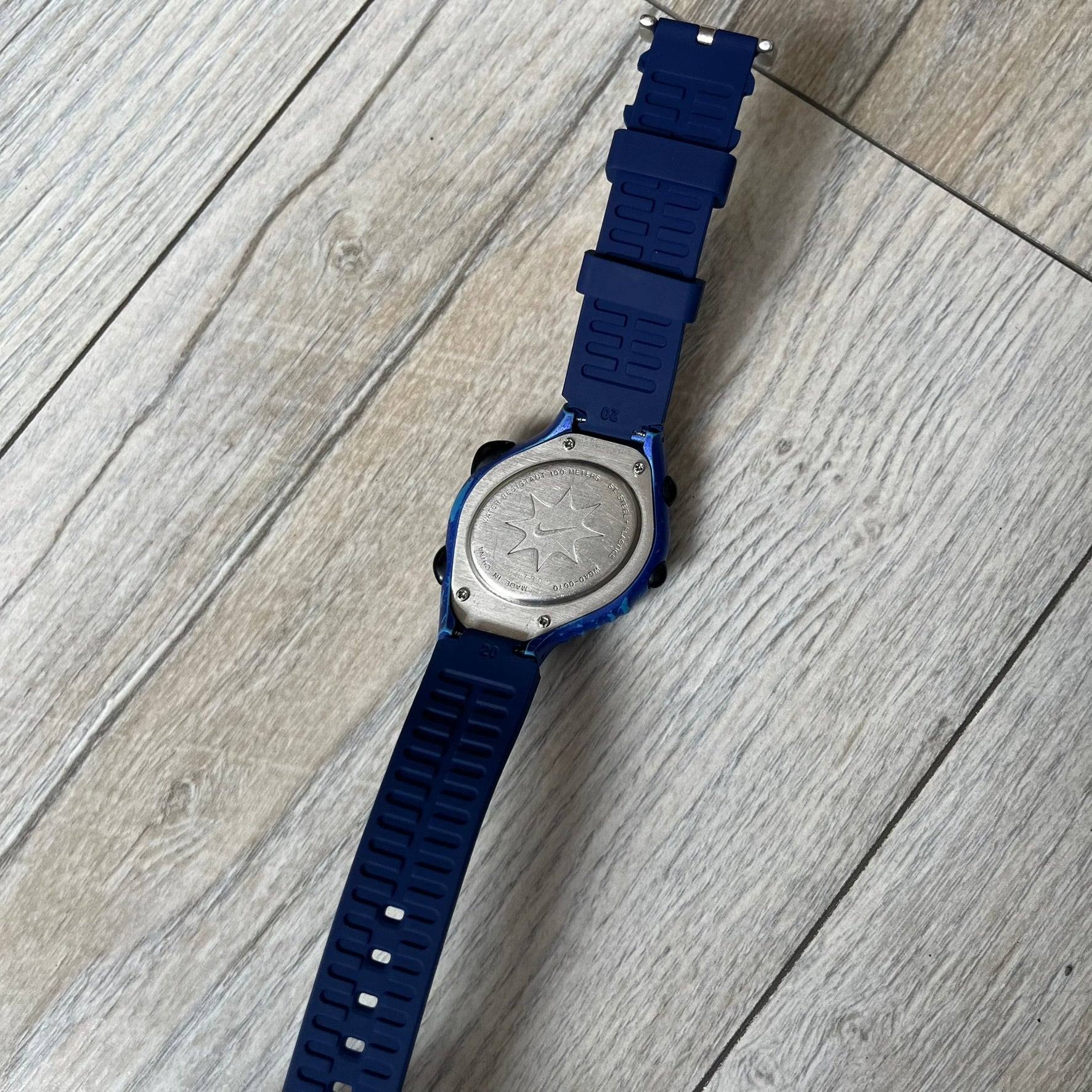 Nike Triax 120 WG40-0010 Digital Watch features a blue case with a black Nike Swoosh above the display and a black control button below it. The watch has a black digital screen with light-colored numerals and is fitted with navy-blue replacement straps that are not original Nike straps. Constructed from stainless steel and plastic materials, it offers 100-meter water resistance, 120-lap memory, and multiple timing and training modes.